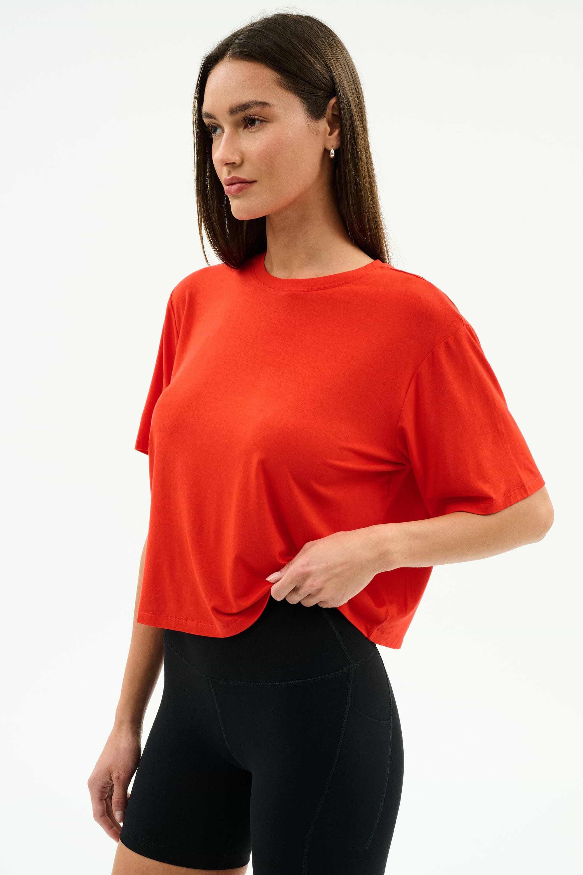 A woman stands against a plain white background, ready for gym workouts in the SPLITS59 Djuna Oversized Jersey Tee in Lotus Red and black fitted shorts.