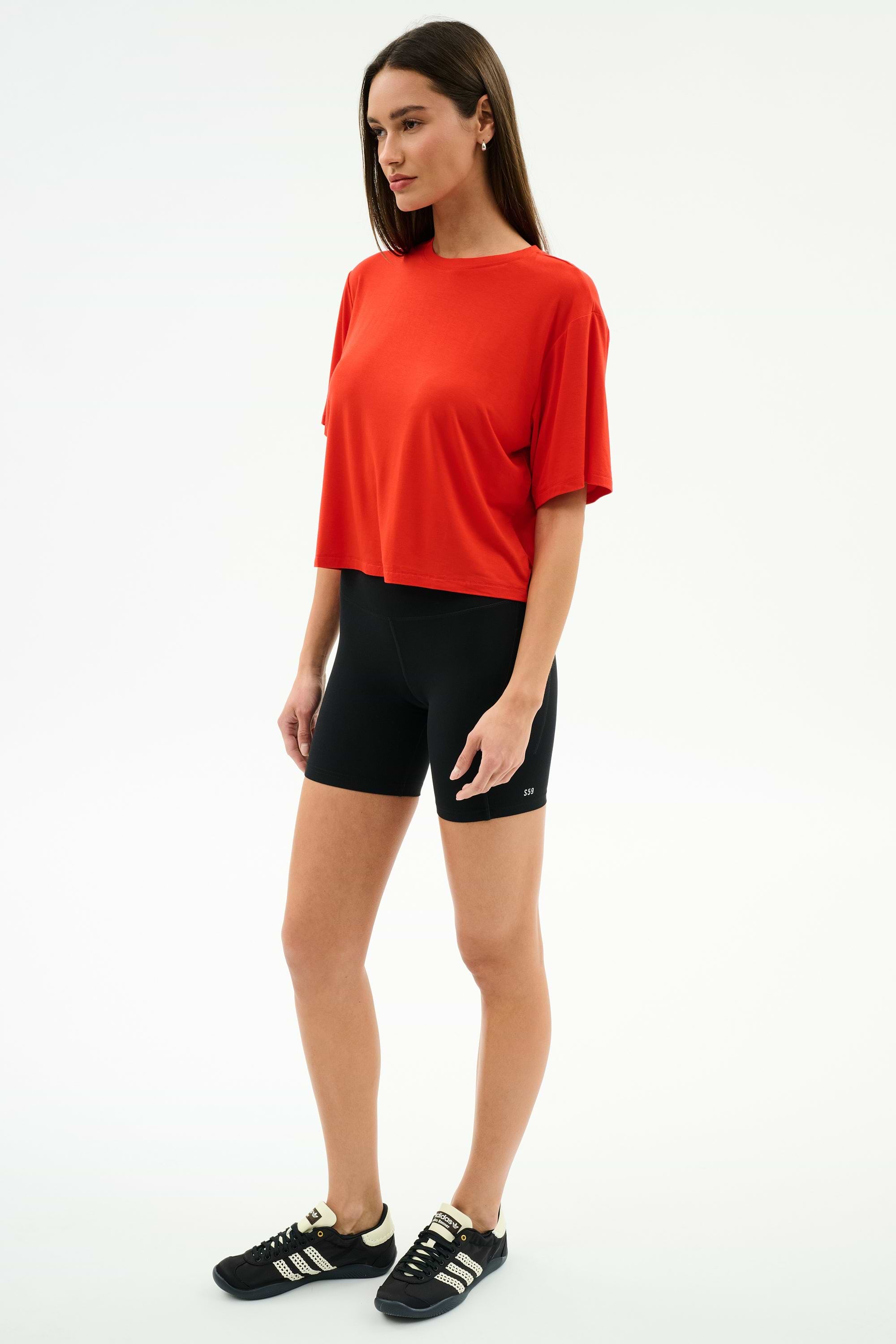 Woman standing against a plain white background, wearing the SPLITS59 Djuna Oversized Jersey Tee in Lotus Red, black bike shorts, and black Adidas sneakers—ideal for gym or outdoor workouts.