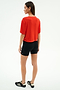 A person stands facing away, wearing the SPLITS59 Djuna Oversized Jersey Tee in Lotus Red, paired with black fitted shorts and black Adidas sneakers against a plain white background—ideal for gym workouts.