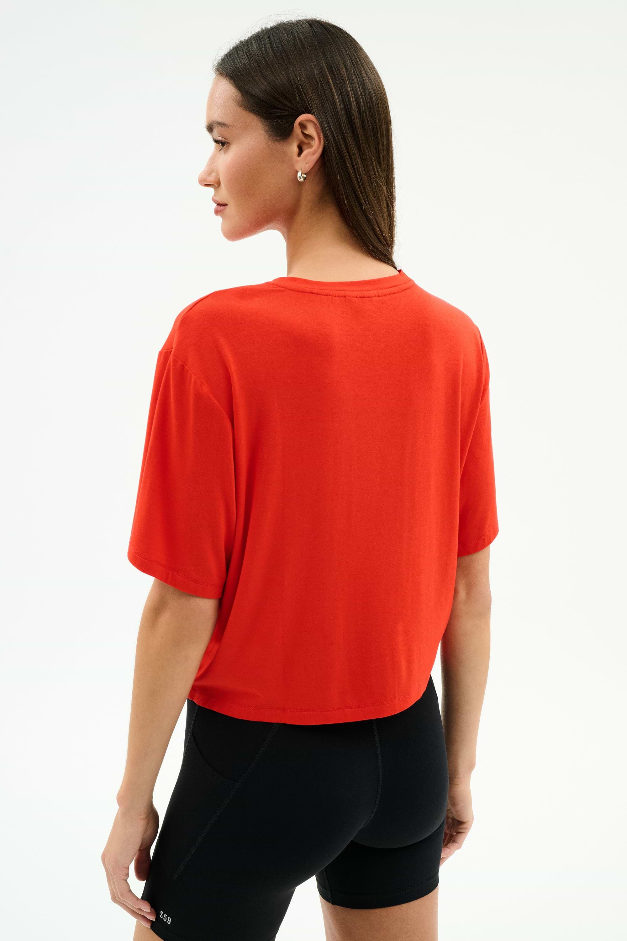A woman stands facing slightly away from the camera, wearing the SPLITS59 Djuna Oversized Jersey Tee in Lotus Red and black athletic shorts, perfect for gym workouts, against a plain white background.