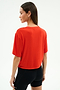 A woman stands facing slightly away from the camera, wearing the SPLITS59 Djuna Oversized Jersey Tee in Lotus Red and black athletic shorts, perfect for gym workouts, against a plain white background.