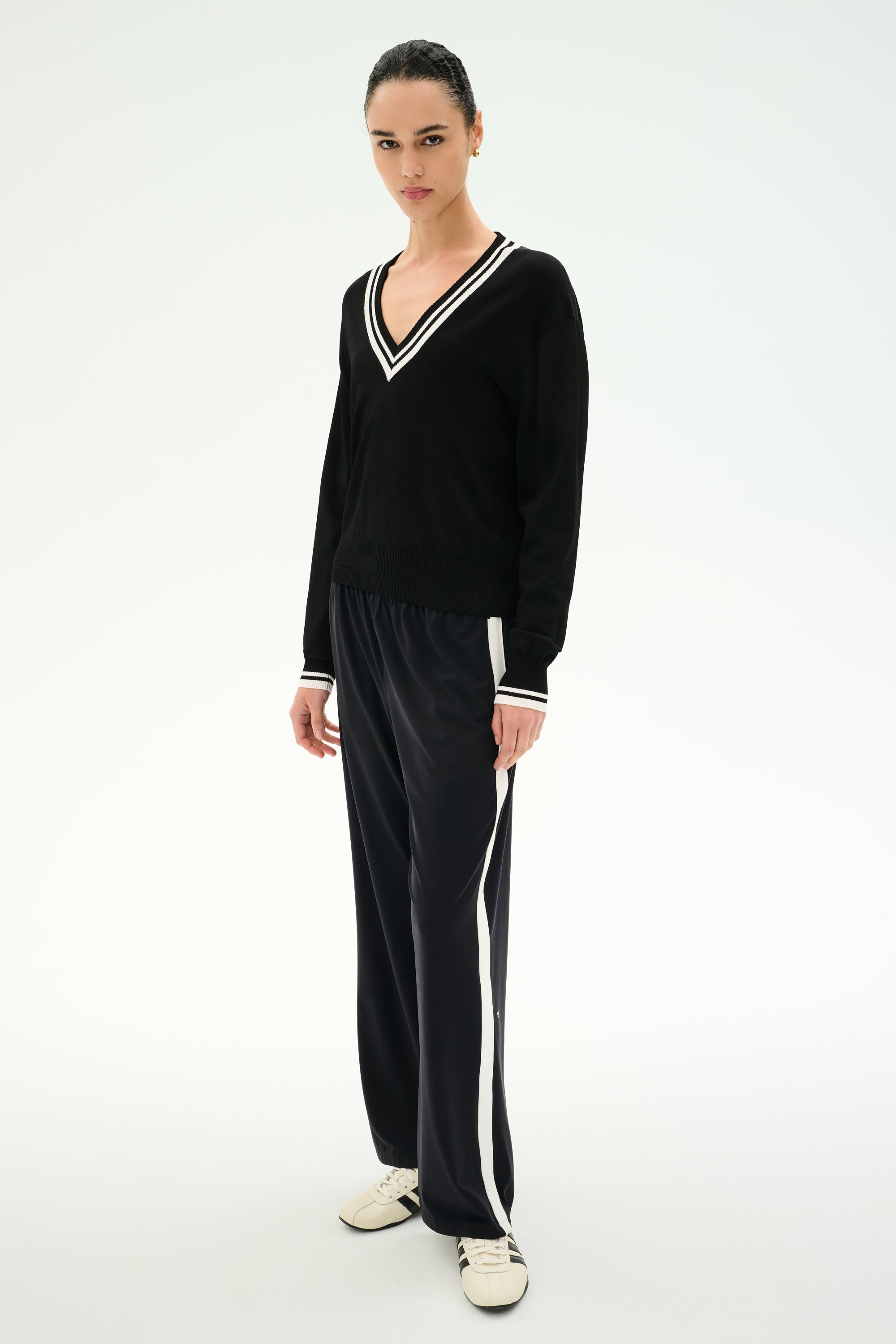 Wearing the SPLITS59 Jemma Knit V Neck in Black/White, this person pairs the soft rayon sweater featuring white trim with black wide-leg pants with side stripes and white sneakers, standing against a plain light background.