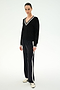 Wearing the SPLITS59 Jemma Knit V Neck in Black/White, this person pairs the soft rayon sweater featuring white trim with black wide-leg pants with side stripes and white sneakers, standing against a plain light background.