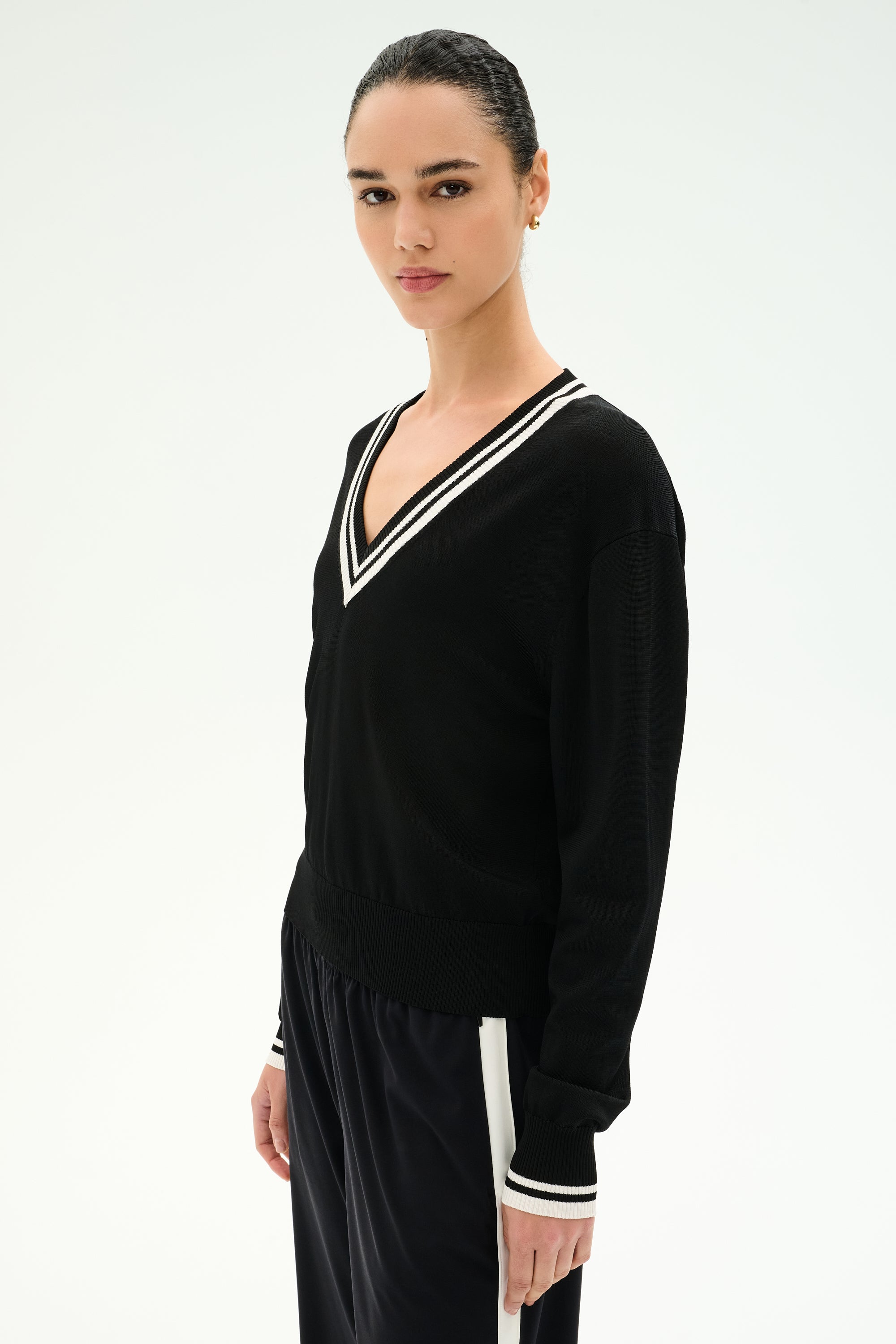 A woman stands against a plain, light background wearing the SPLITS59 Jemma Knit V Neck in Black/White—a soft rayon-nylon sweater featuring a white-trimmed V-neck—paired with matching black pants accented by a white stripe.