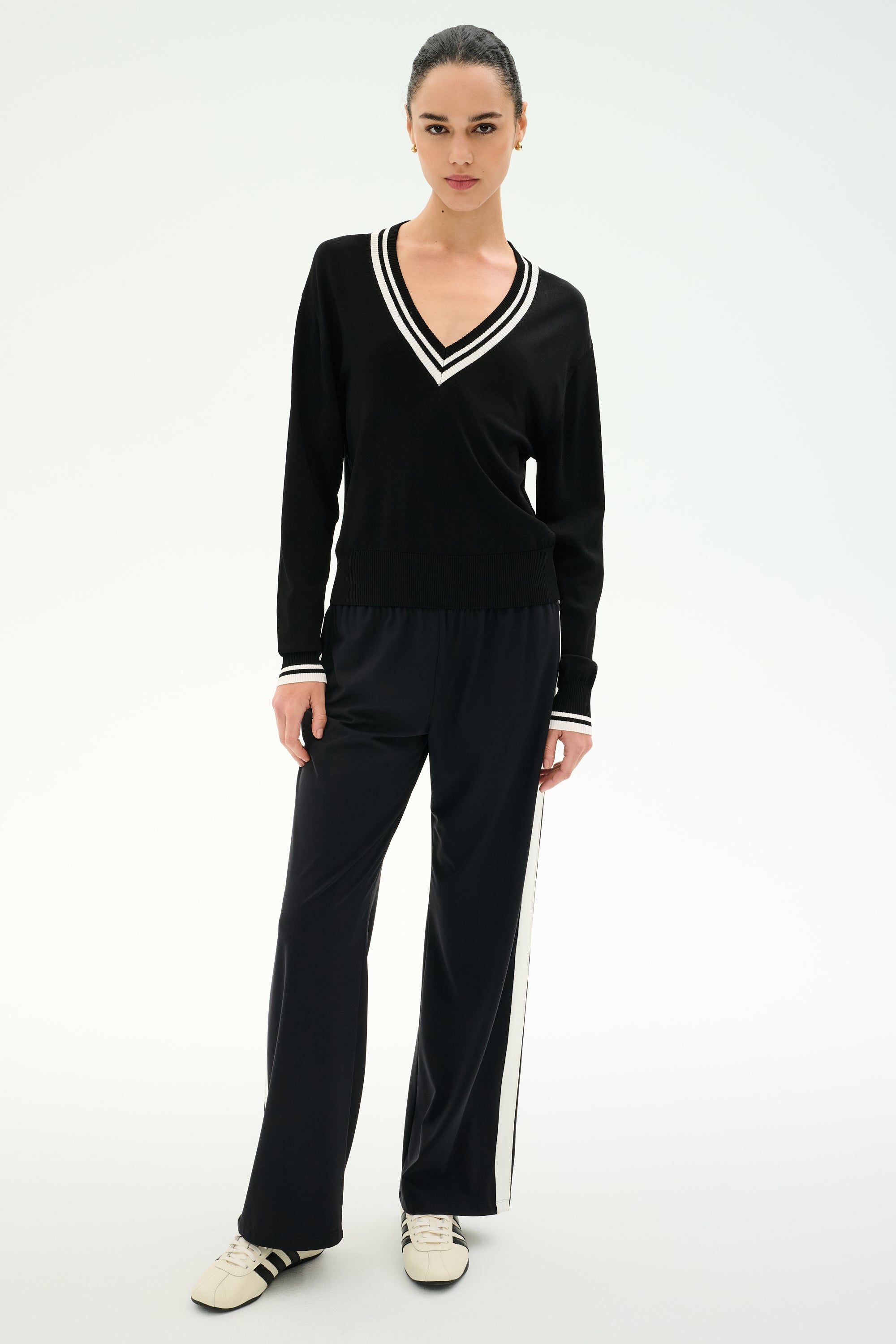 A person stands against a plain background wearing the SPLITS59 Jemma Knit V Neck in Black/White, expertly crafted from a soft rayon-nylon blend, paired with black wide-leg pants featuring a white side stripe and crisp white sneakers.