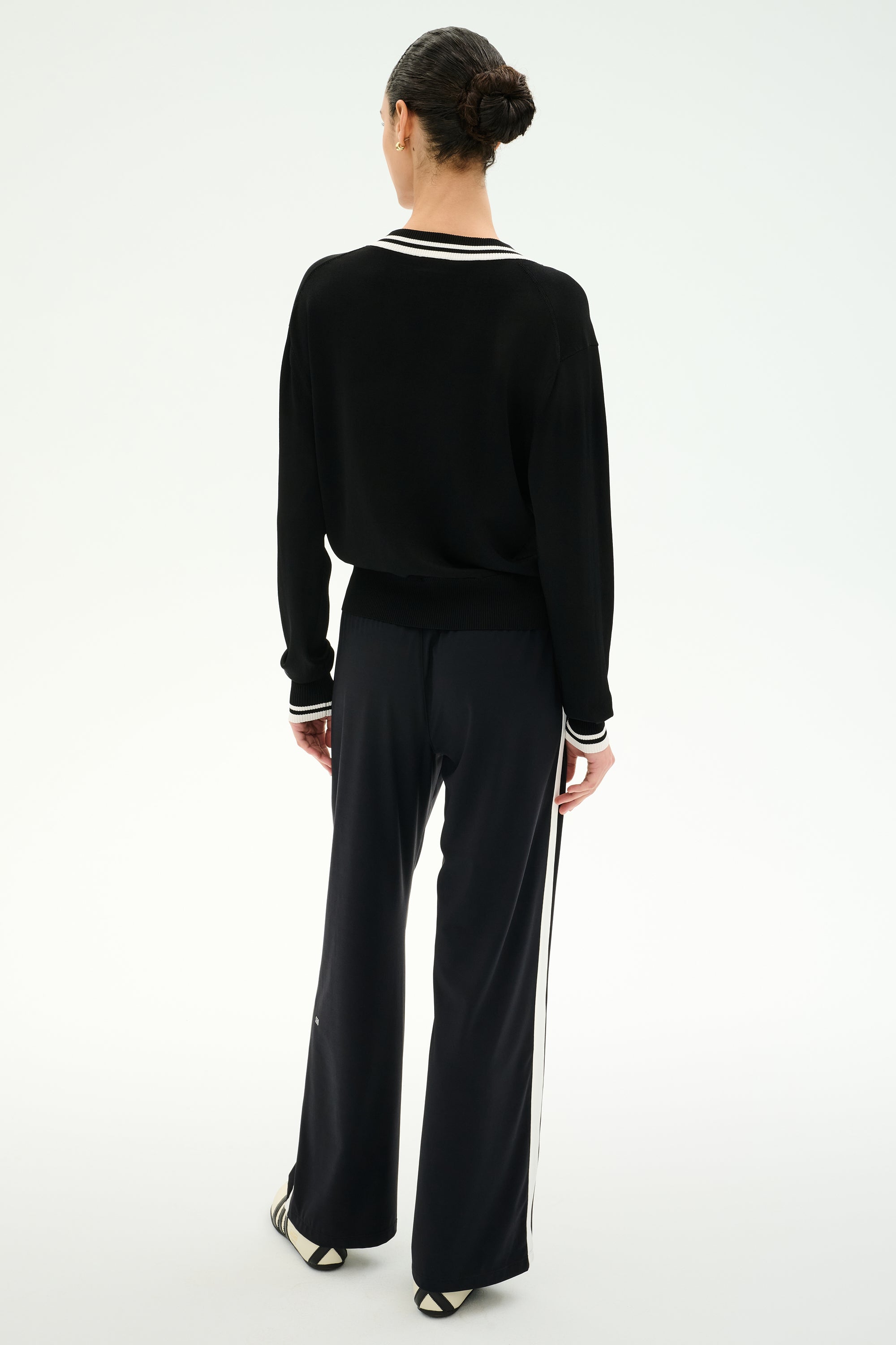 A person with dark hair in a bun stands facing away, wearing the SPLITS59 Jemma Knit V Neck in Black/White—a long-sleeve top paired with wide-leg black pants featuring white side stripes, all crafted from soft rayon fabric—against a plain white background.