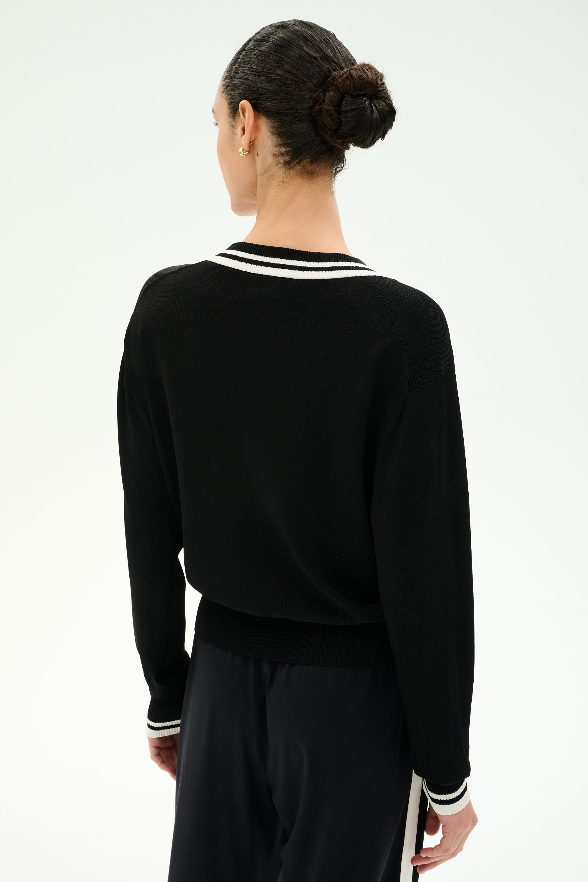 A person stands facing away, wearing the SPLITS59 Jemma Knit V Neck in Black/White paired with black soft rayon pants. Their hair is tied back in a neat bun, and the background is plain white.