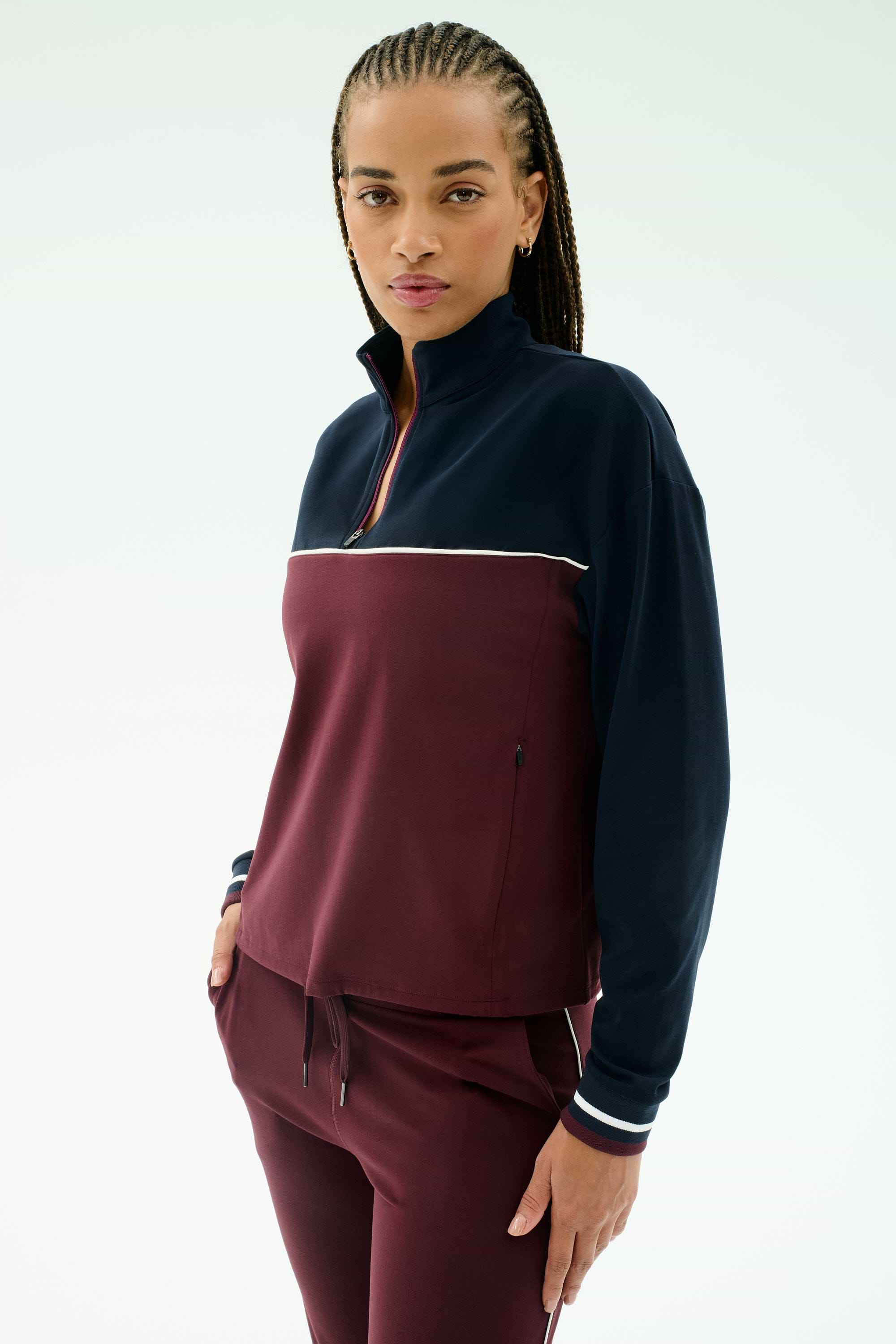 A woman wears the SPLITS59 Millie Airweight Half Zip in Dark Cherry/Indigo, a comfortable nylon-spandex blend track suit, standing with one hand in her pocket and looking at the camera against a plain white background.