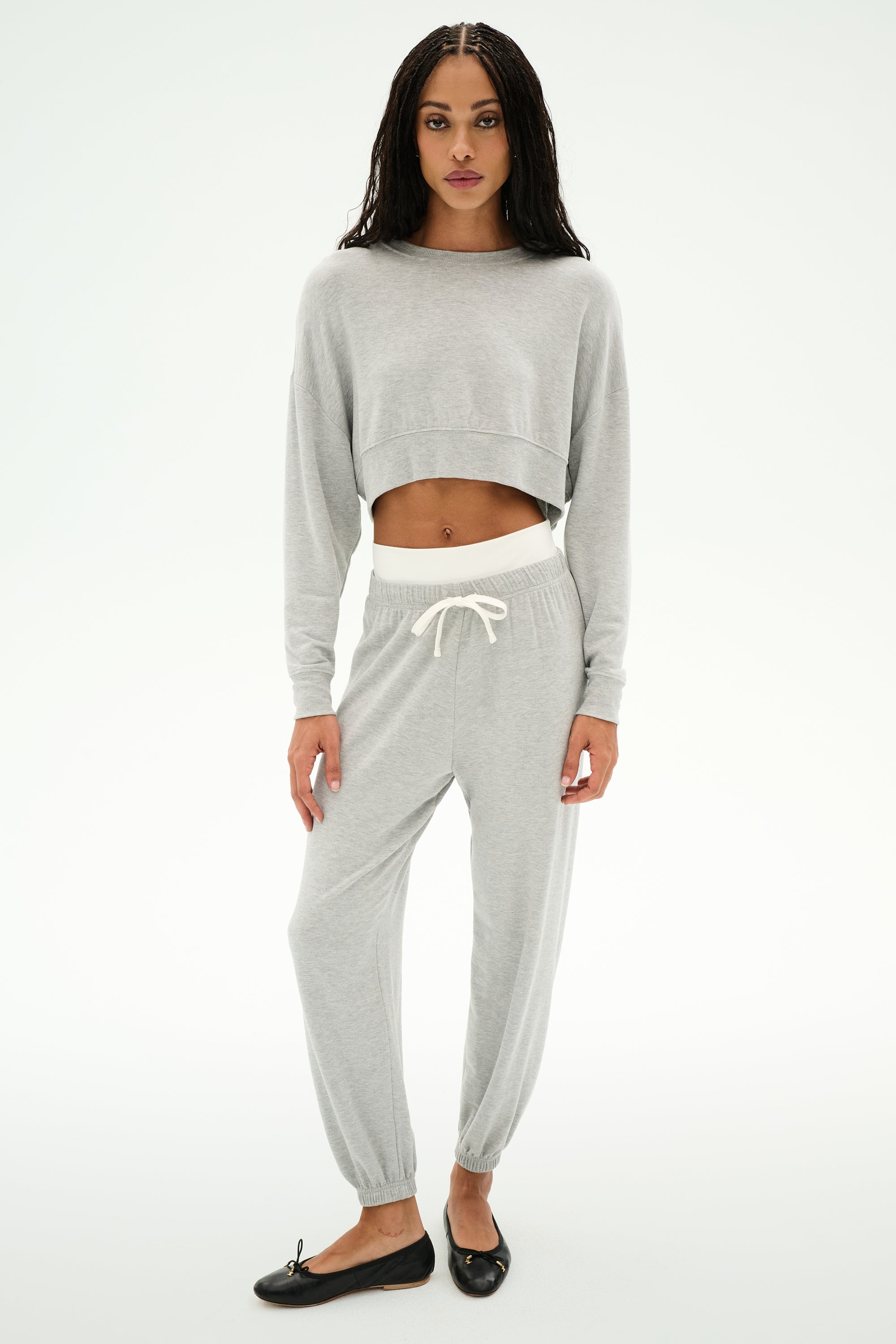 A woman stands against a plain background wearing SPLITS59's Andie Dual Waist Sweatpant in Heather Grey/White—soft modal blend joggers with a drawstring—paired with a light grey cropped sweatshirt and black ballet flats.