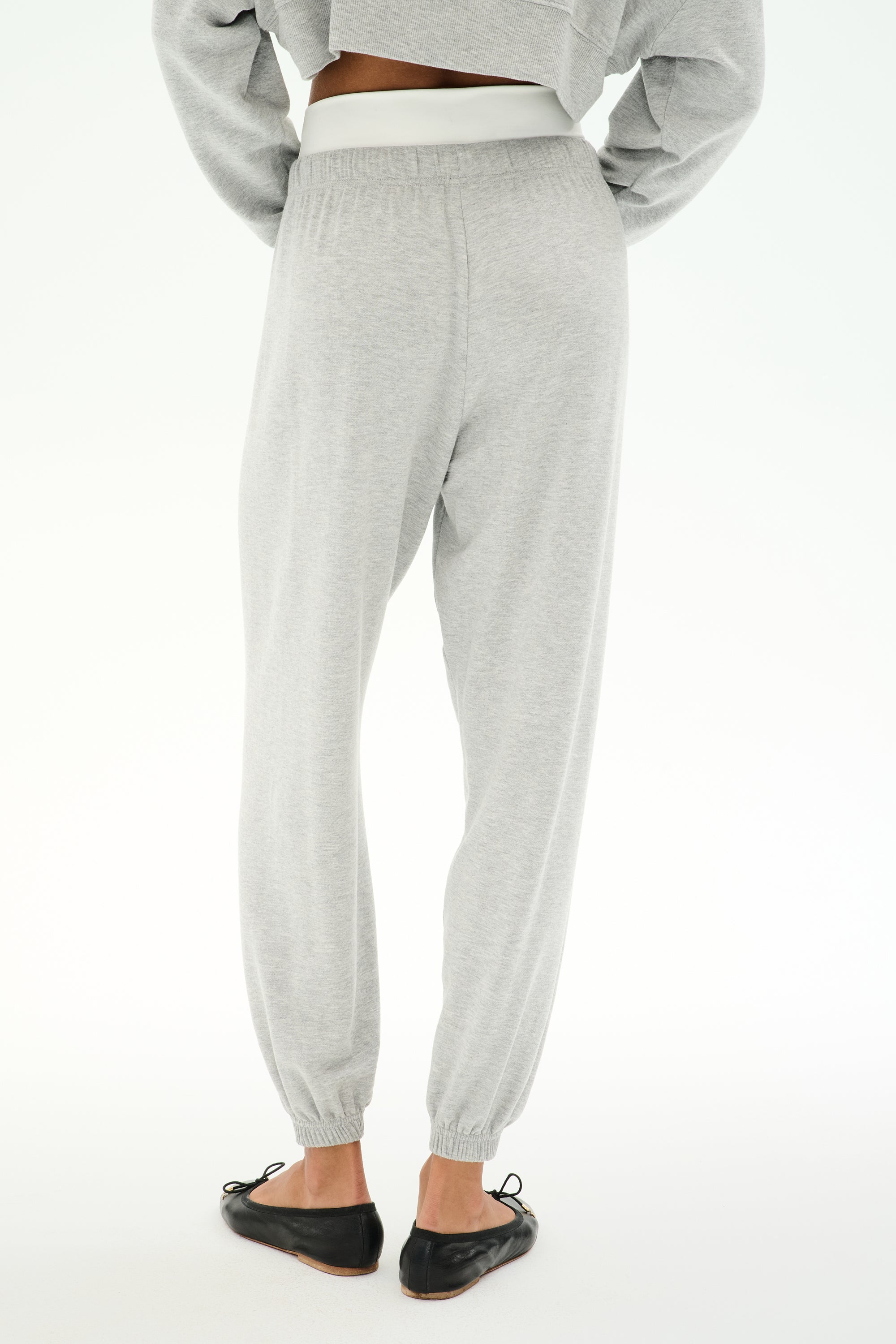 A person wears SPLITS59's Andie Dual Waist Sweatpant in Heather Grey/White with an elastic waist and cuffs, paired with black flats and a cropped gray long-sleeve top, standing on a white background.