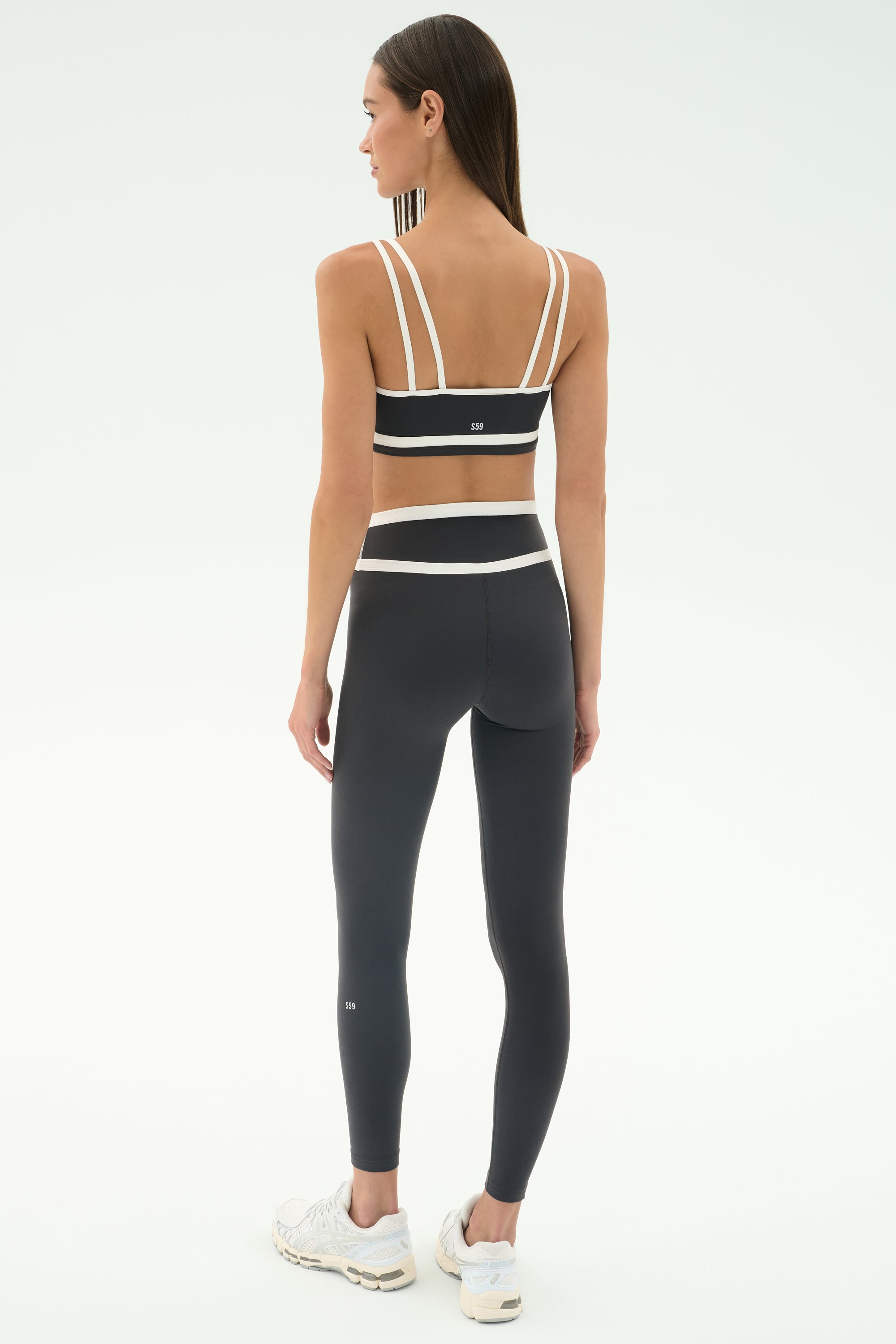 A woman faces away in the SPLITS59 Eva Rigor Double Strap Bra in Graphite/White, paired with high-waisted leggings and white sneakers—an ideal gym-ready look—against a plain white background.