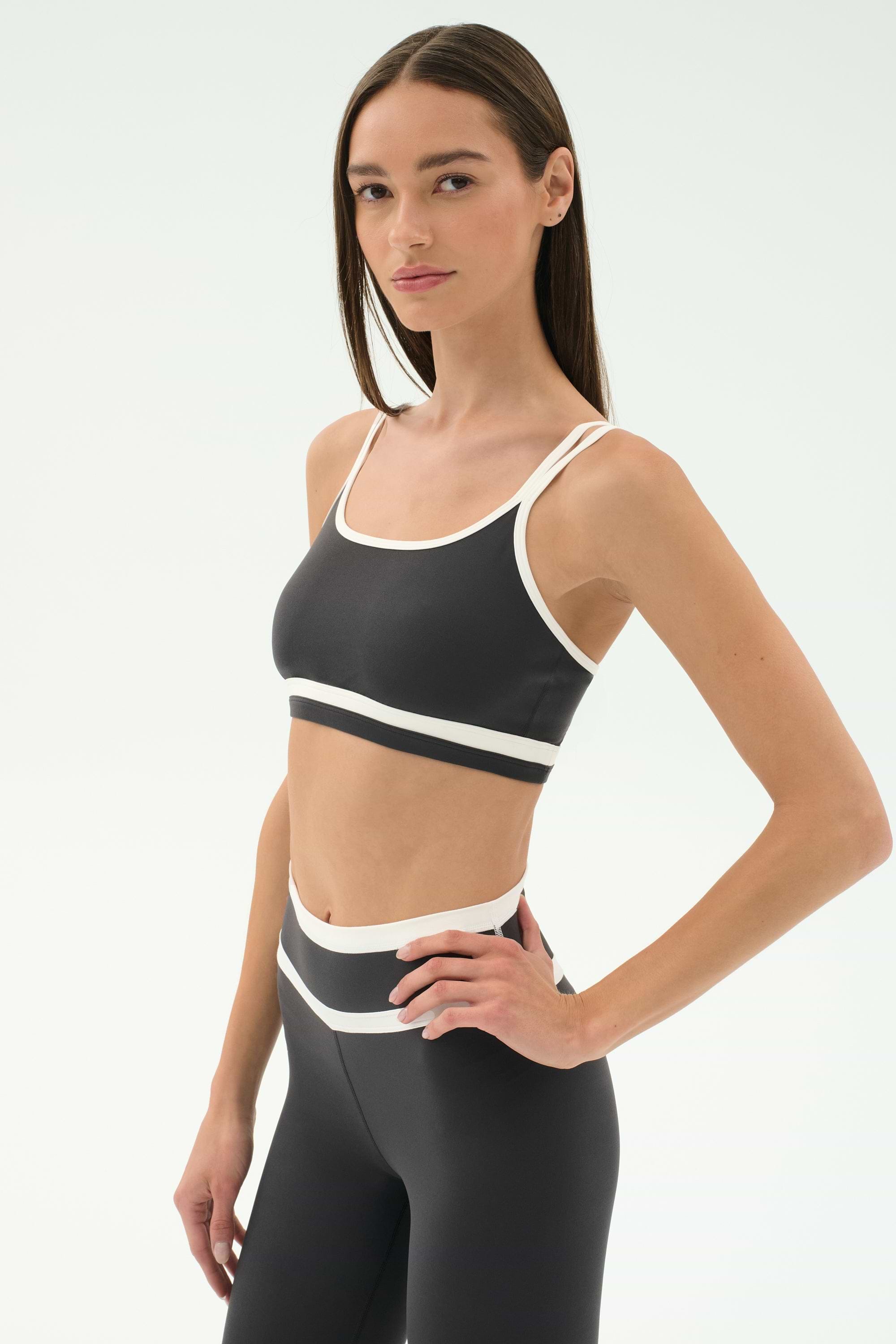 A woman wears the SPLITS59 Eva Rigor Double Strap Bra in Graphite/White with matching high-waisted leggings, ideal for gym workouts, posing against a plain white background.