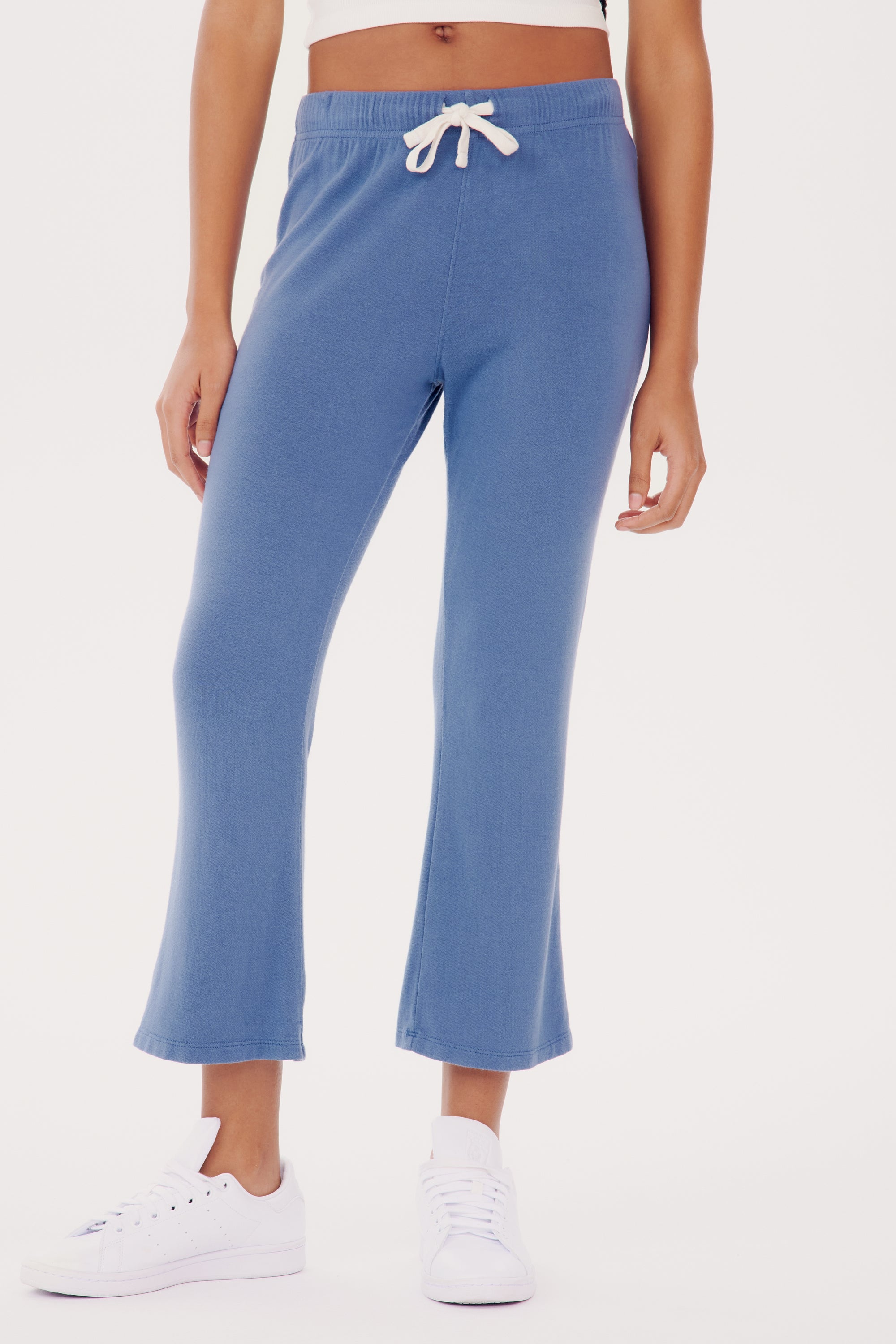 Woman standing in SPLITS59 Brooks Fleece Cropped Flare - Steel Blue sweatpants and white sneakers, cropped view showing only from waist to shoes.
