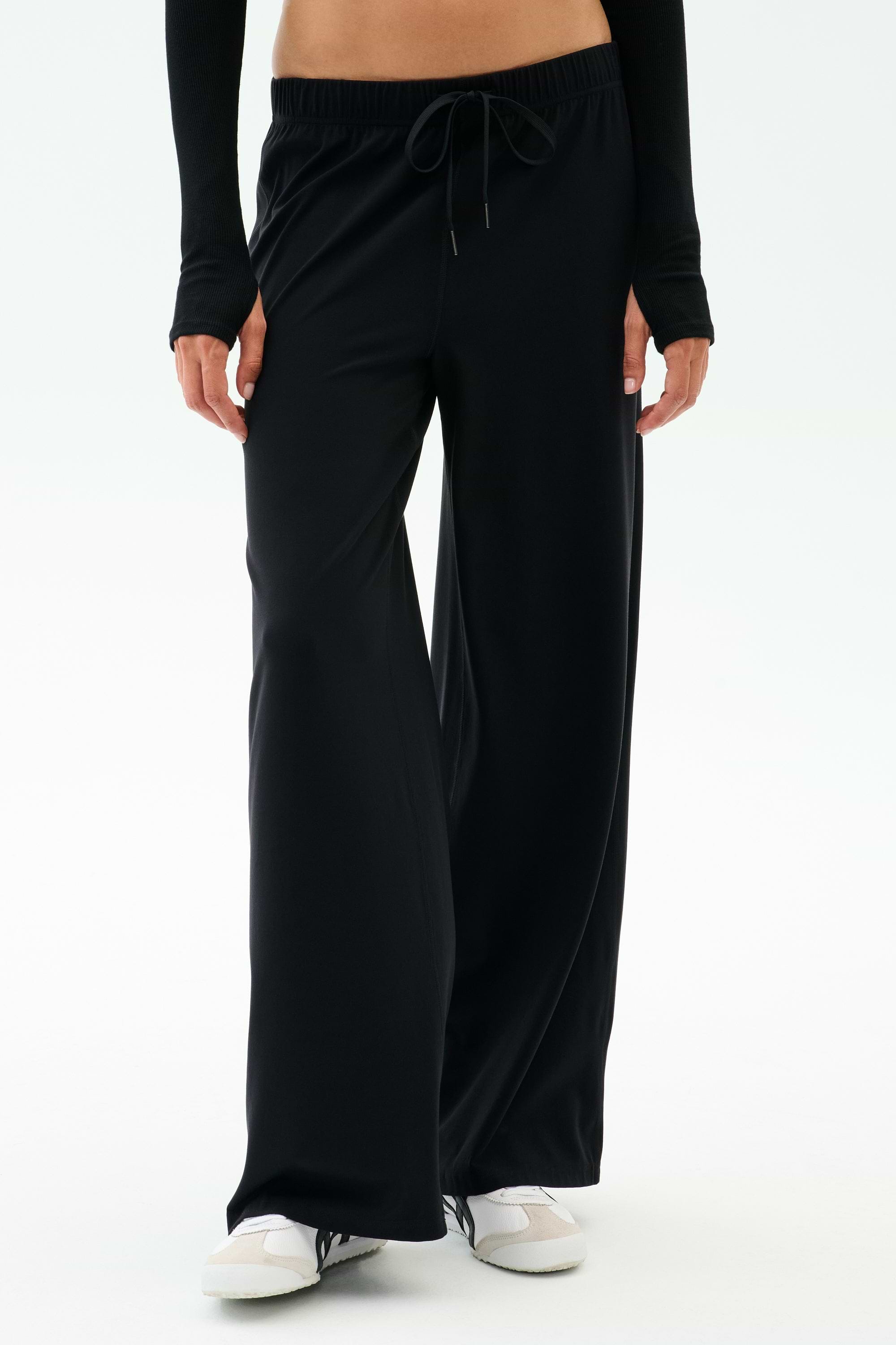 Wearing SPLITS59's Brynn Airweight Pant in Black with a black long-sleeve top and white sneakers, the person stands against a plain white background.