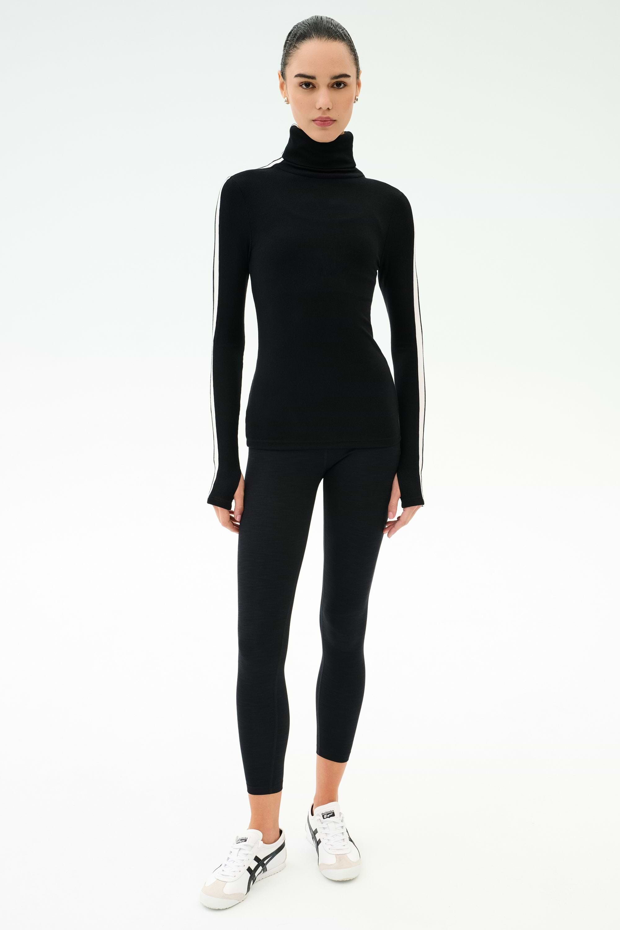 A woman faces forward in a SPLITS59 Jackson Rib Full Length Turtleneck - Black/White, paired with black leggings and white sneakers, set against a plain white background.