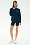 A woman stands against a plain background wearing the SPLITS59 Joy Velour Crewneck in Indigo, navy cycling shorts, white socks, and white athletic shoes.