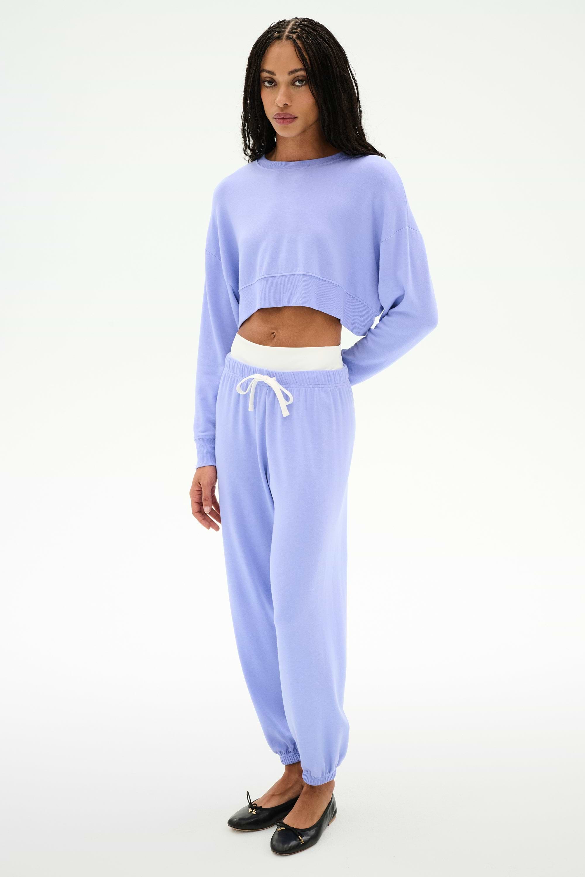 A person wears the SPLITS59 Andie Dual Waist Sweatpant in Purple Haze/White, featuring a stretchy fit, paired with a lavender cropped sweatshirt, standing against a plain white background.