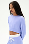 A woman with long braided hair wears the SPLITS59 Noah Fleece Crop Sweatshirt in Purple Haze, featuring a stretchy fit, paired with matching sweatpants, standing against a plain white background.