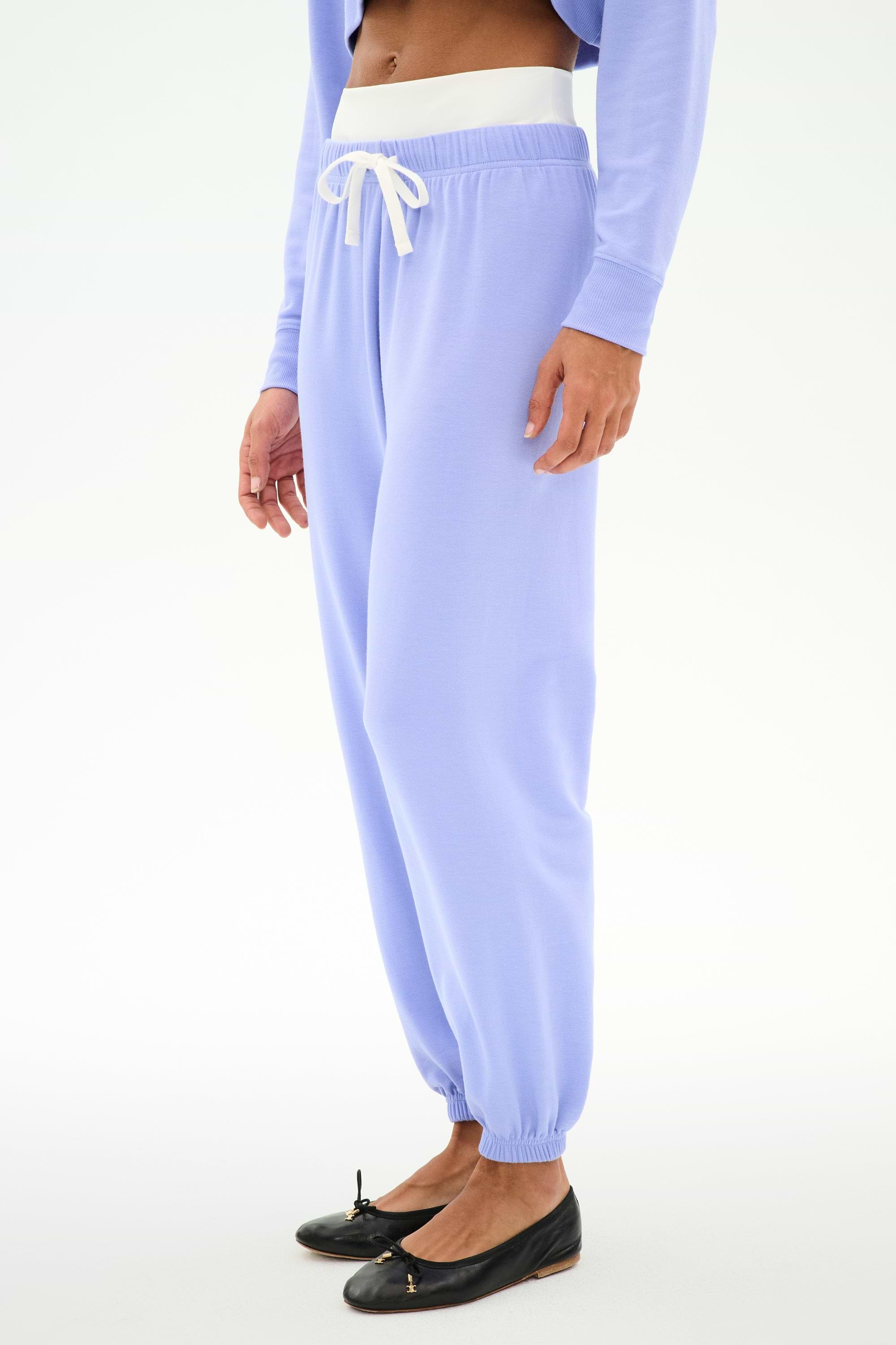 A person wears SPLITS59's Andie Dual Waist Sweatpant in Purple Haze/White with a matching long-sleeve top and black ballet flats, standing against a plain white background.