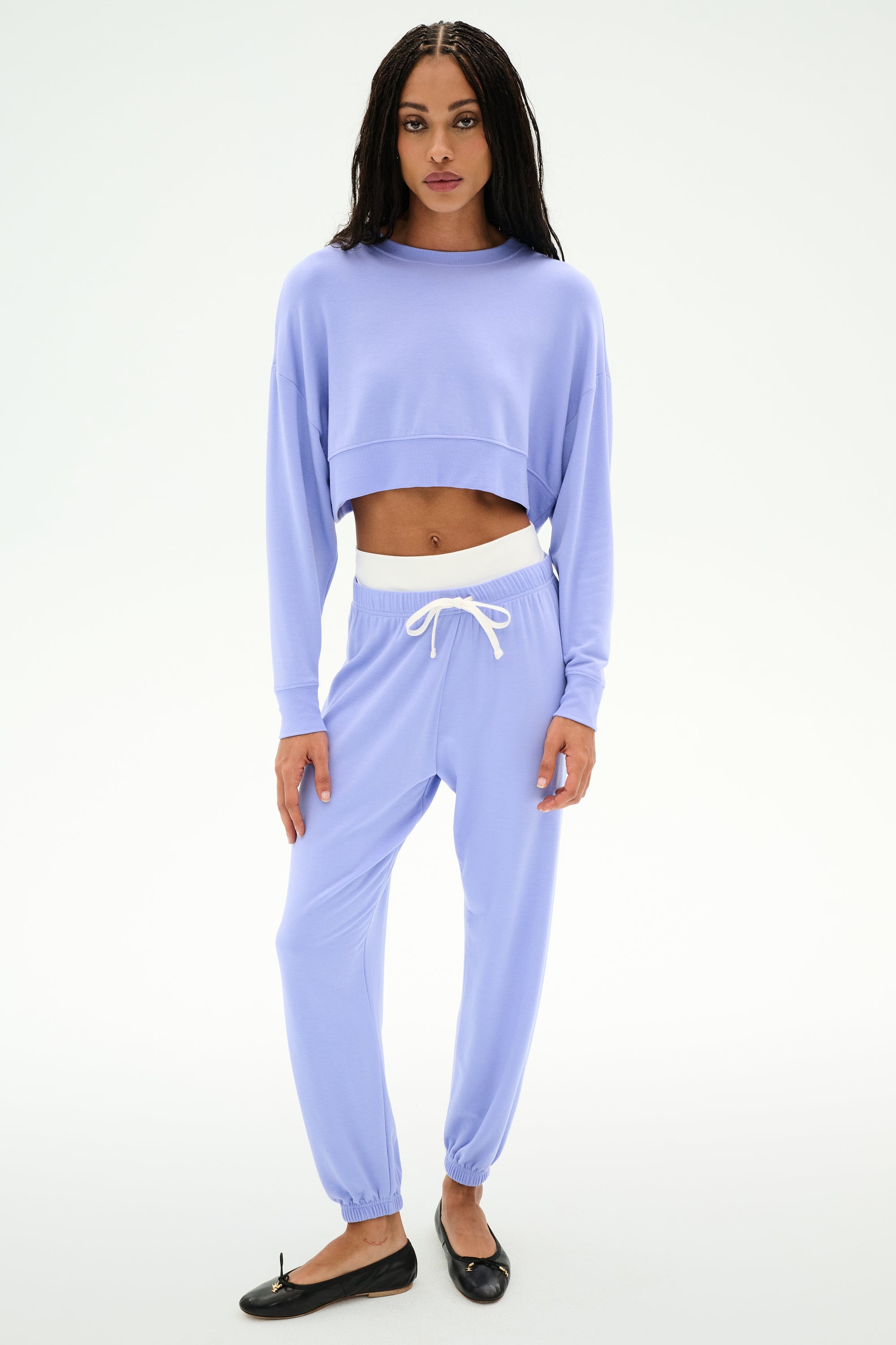 A person stands facing forward, wearing SPLITS59's Andie Dual Waist Sweatpant in Purple Haze/White, crafted from soft modal fabric and paired with a matching light purple long-sleeve cropped top and black flats.