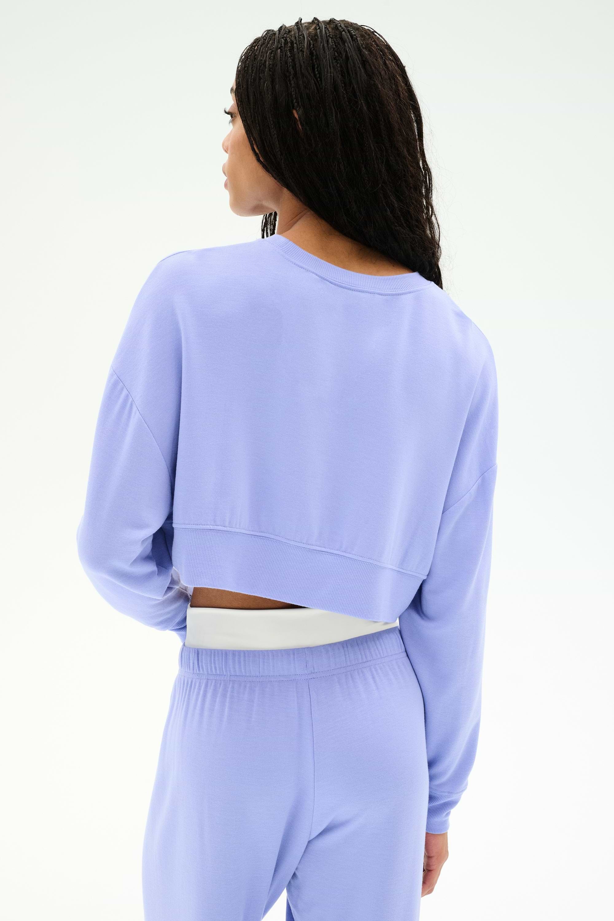 A person with long braided hair is shown from the back wearing the SPLITS59 Noah Fleece Crop Sweatshirt in Purple Haze, paired with matching stretchy pants, set against a plain light background.