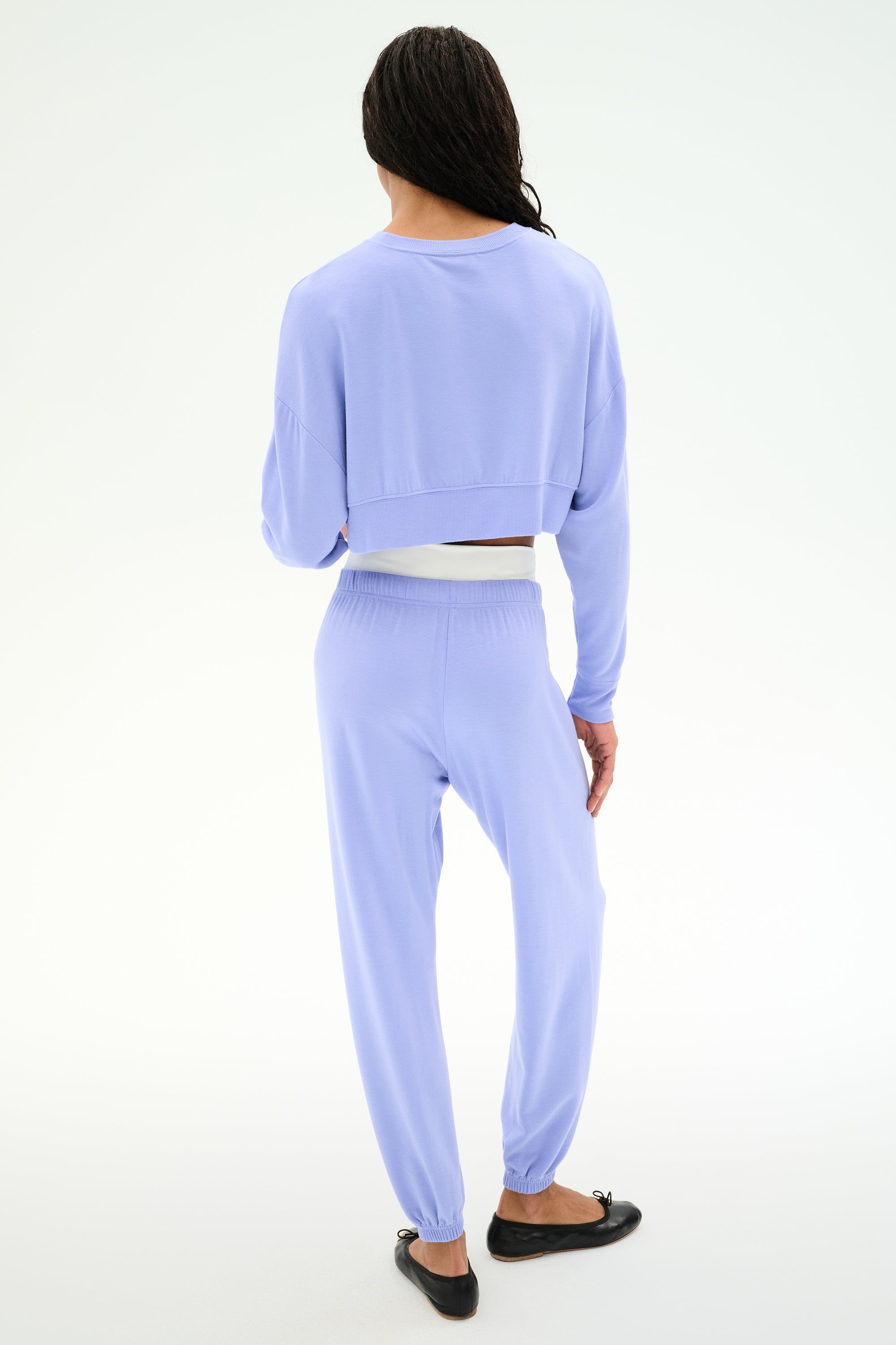 A person stands with their back to the camera, wearing SPLITS59's Andie Dual Waist Sweatpant in Purple Haze/White and a matching light purple long-sleeve top, both crafted from soft modal fabric, paired with black flats.
