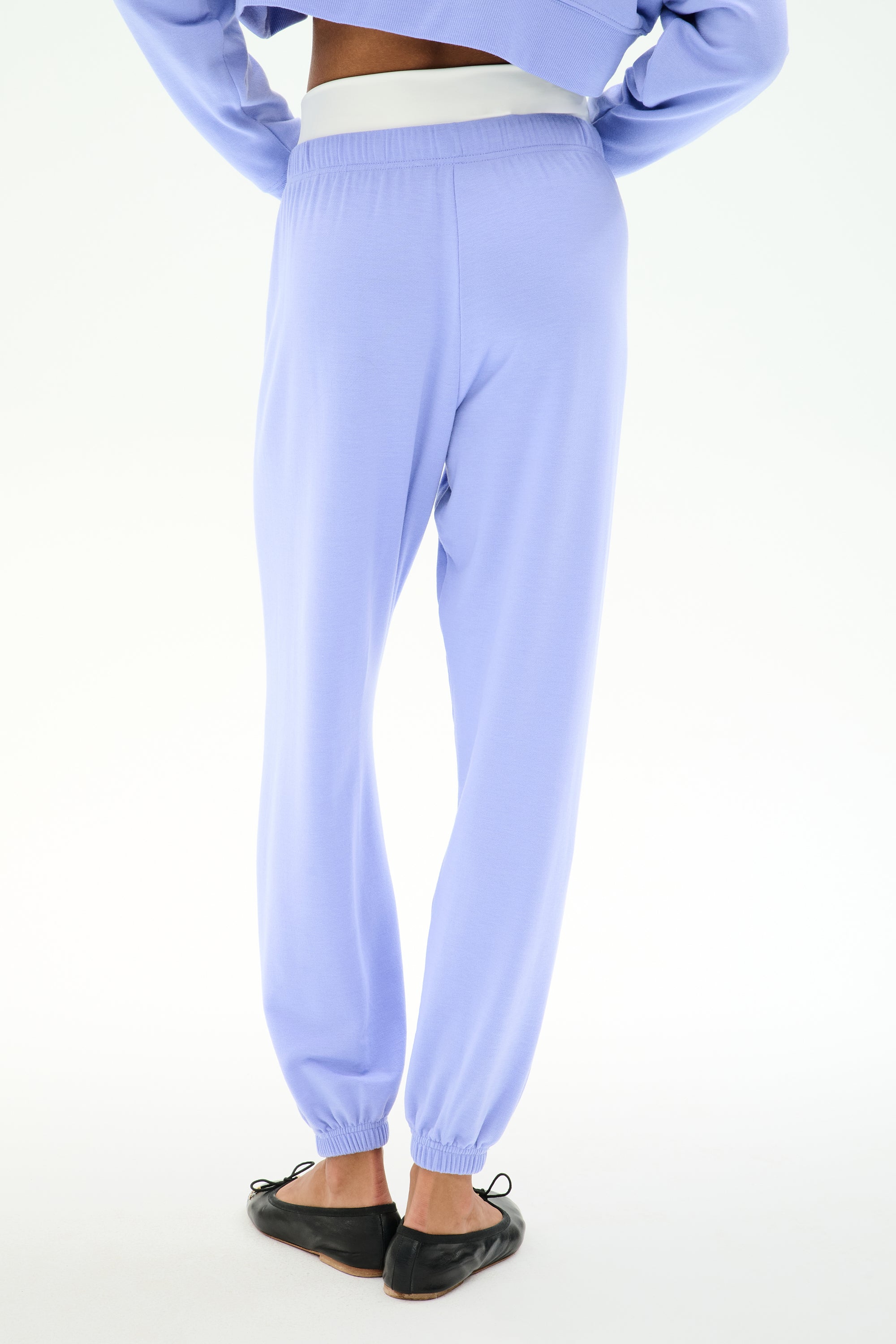 Model showcases the SPLITS59 Andie Dual Waist Sweatpant in Purple Haze/White from the back, featuring a stretchy fit and dual-tone waistband, styled with black slip-on shoes against a white background.