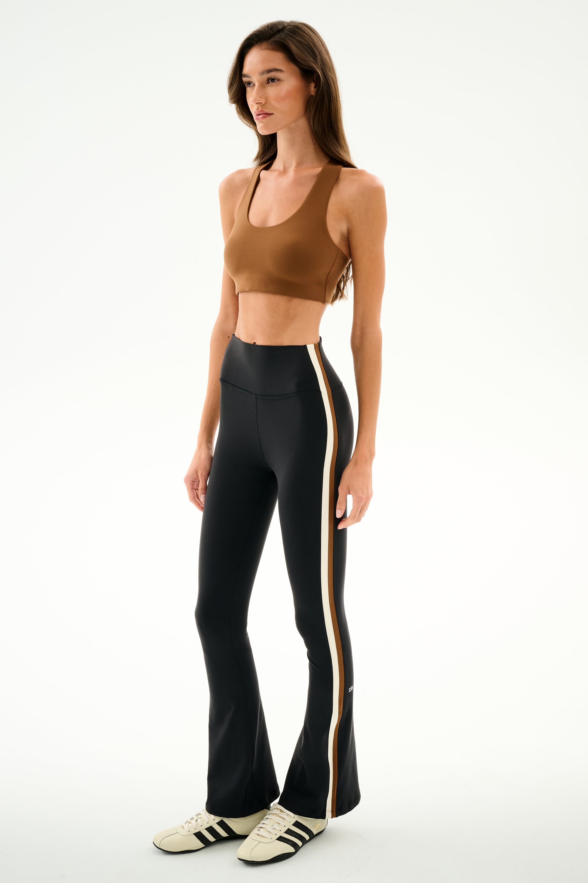 Woman wearing a brown spandex sports bra, SPLITS59 Raquel High Waist Supplex Flare - Black/Creme pants, and white Adidas sneakers against a plain white background.