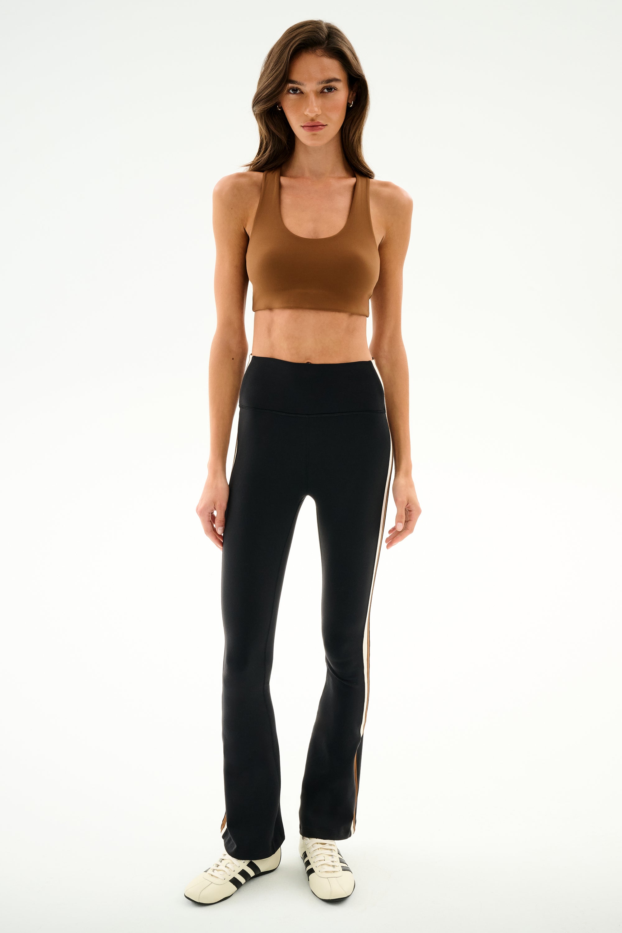Woman in a brown sports bra, white sneakers, and SPLITS59 Raquel High Waist Supplex Flare - Black/Creme against a white background.