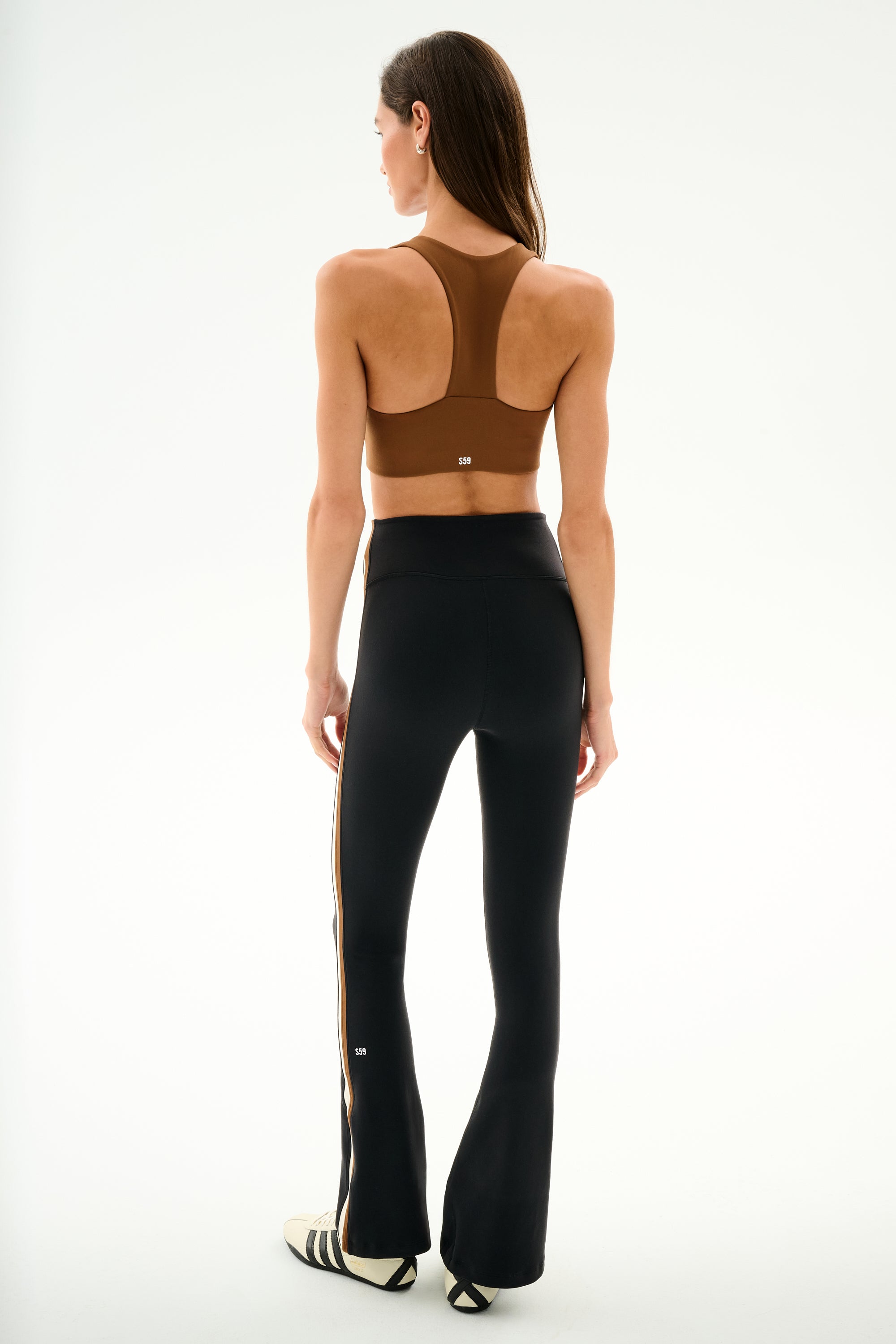 A woman faces away, wearing a brown nylon sports bra, SPLITS59 Raquel High Waist Supplex Flare - Black/Creme pants, and white sneakers with black stripes on a plain white background.