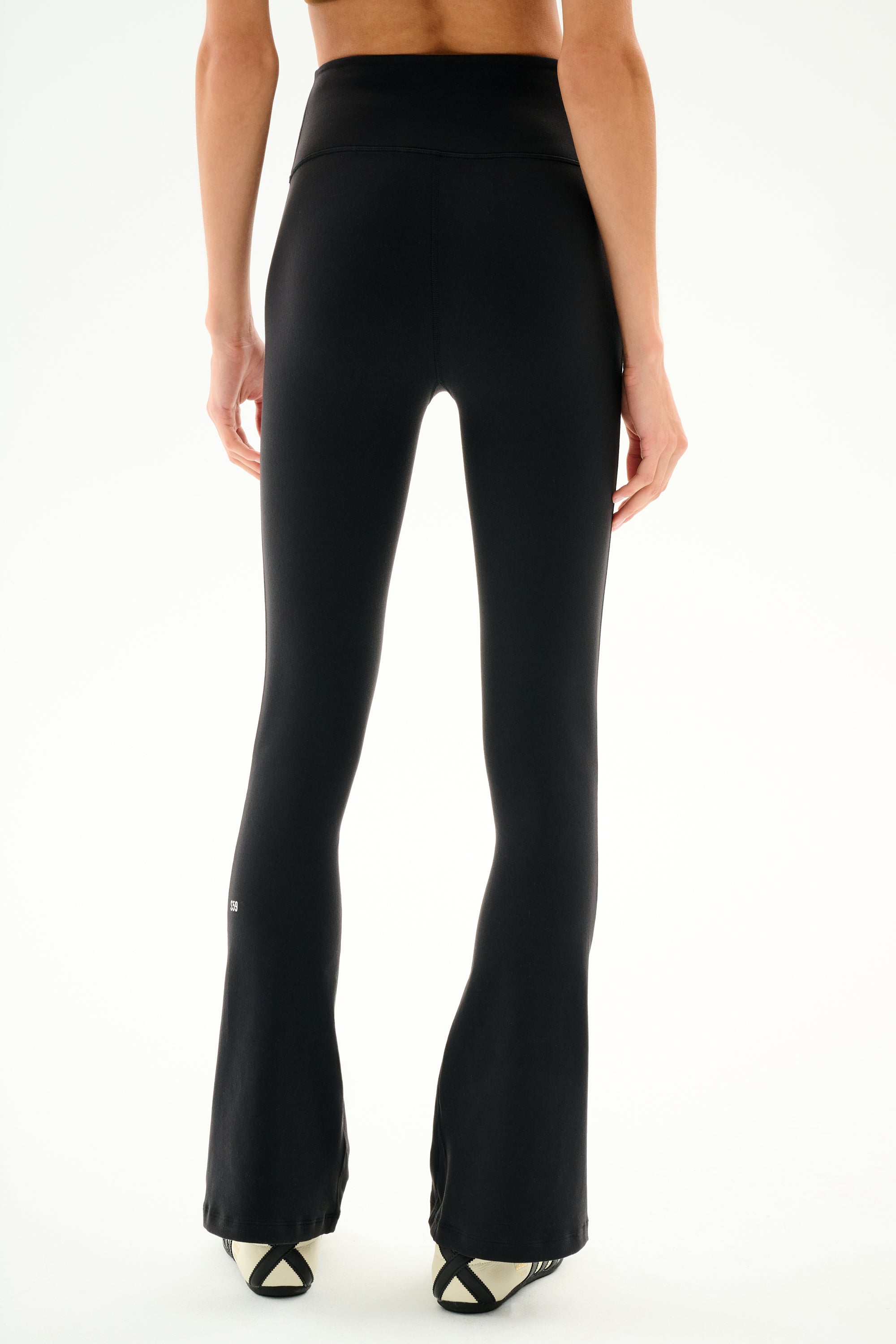 Model wearing SPLITS59 Raquel High Waist Supplex Flare - Black/Creme pants and black-and-white shoes, seen from the back against a plain white background.