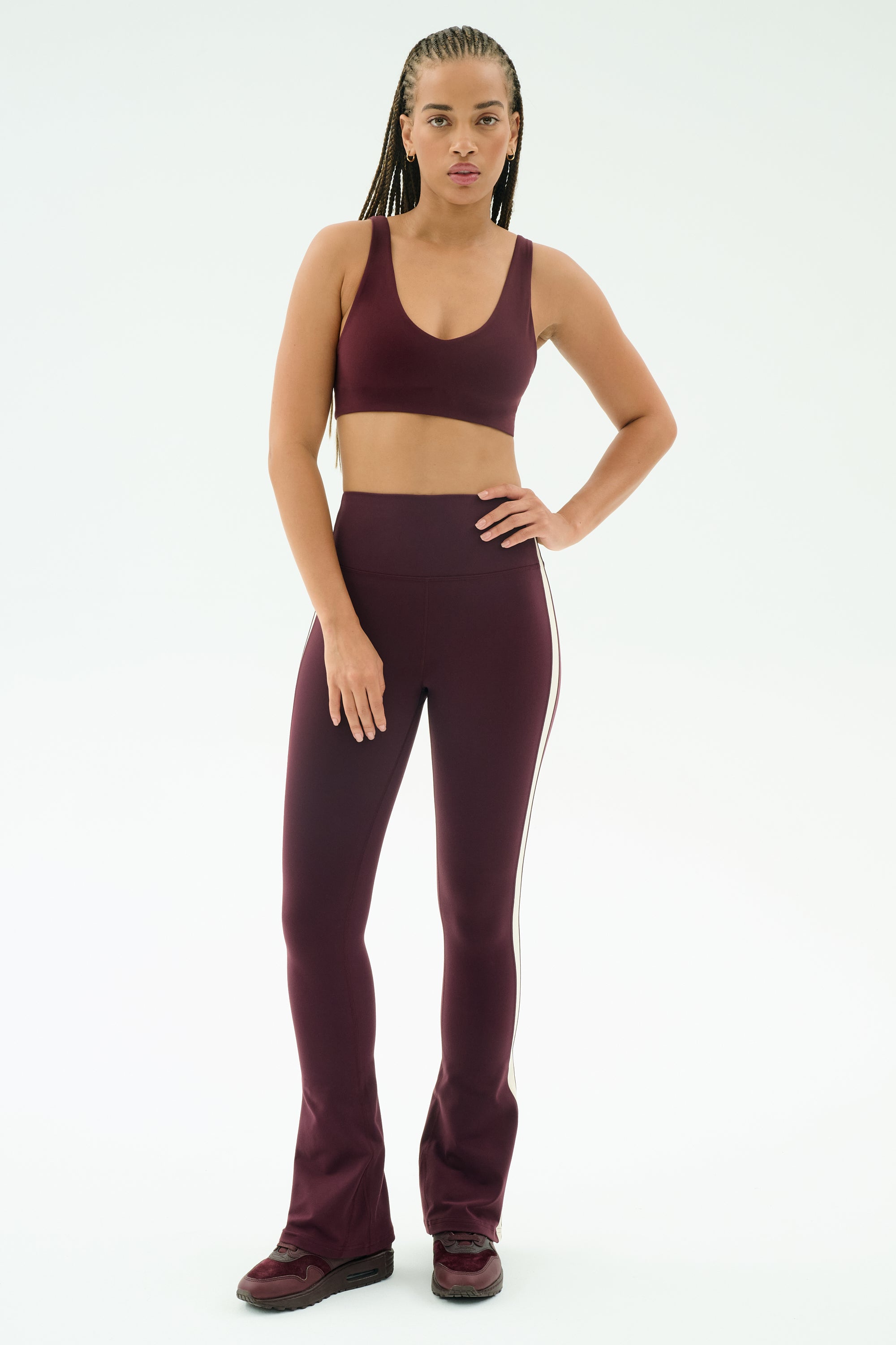 A person stands facing forward, wearing SPLITS59's Raquel High Waist Flared Legging in Dark Cherry/Creme with a matching sports bra and sneakers, set against a plain white background.