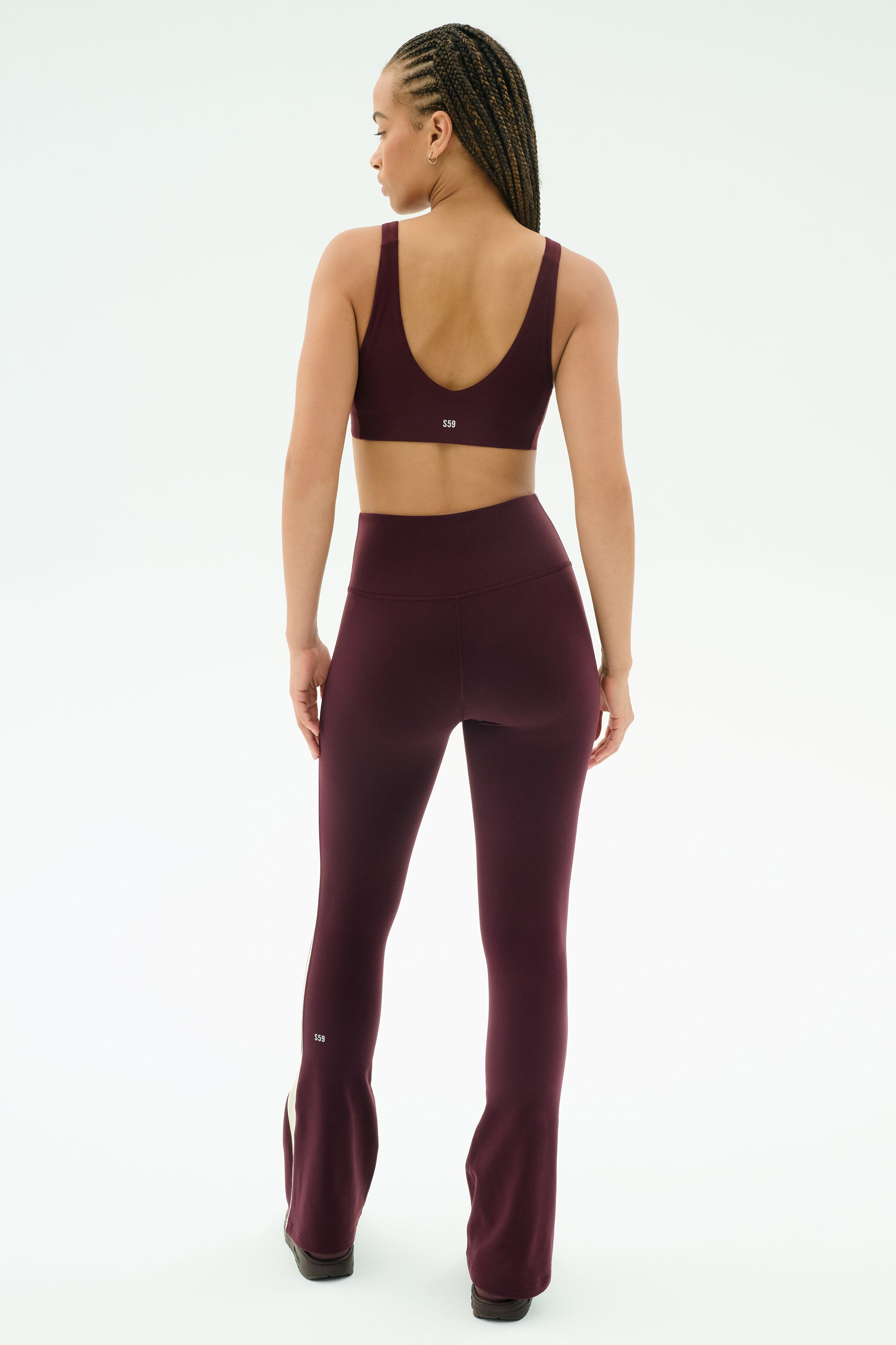 A person with braided hair faces away, wearing SPLITS59’s Raquel High Waist Flared Legging in Dark Cherry/Creme and a matching spandex sports bra.