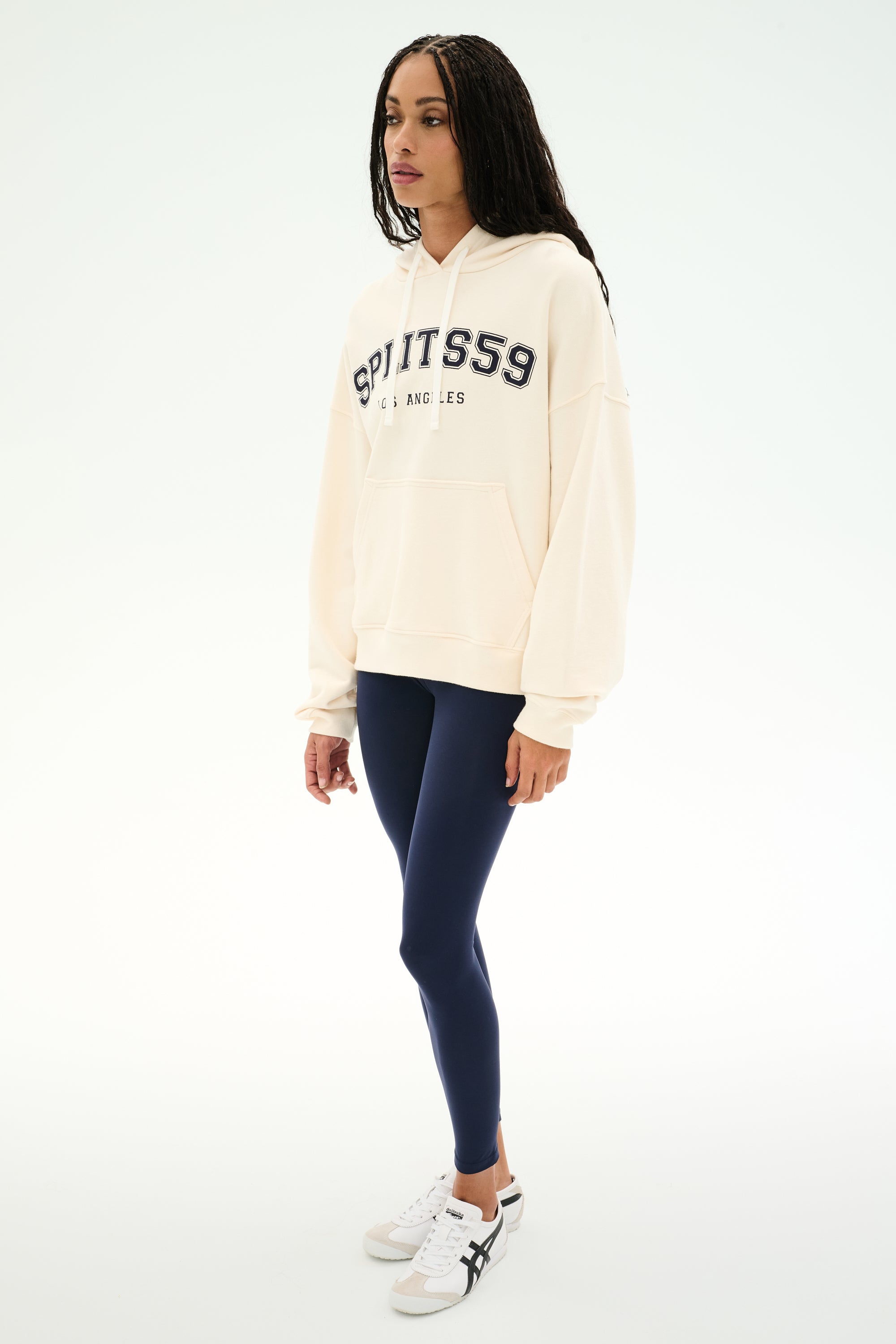 A woman stands against a plain white background wearing the SPLITS59 Stella French Terry Oversized Hoodie in Creme/Splits59 Los Angeles, paired with navy leggings and white sneakers.