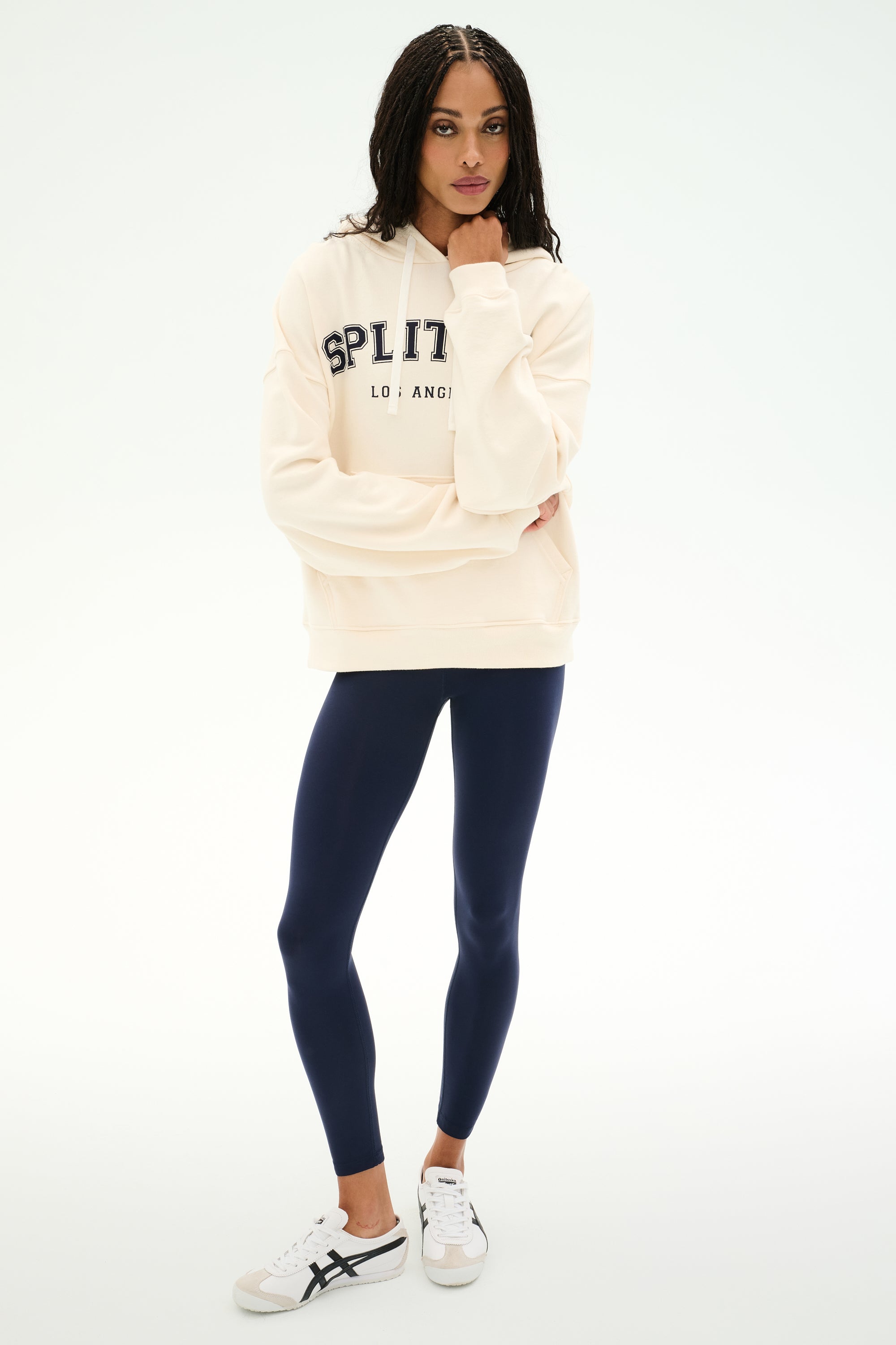 A person stands against a plain white background wearing the SPLITS59 Stella French Terry Oversized Hoodie in creme, featuring "Splits59 Los Angeles" text, paired with navy blue leggings and white sneakers.