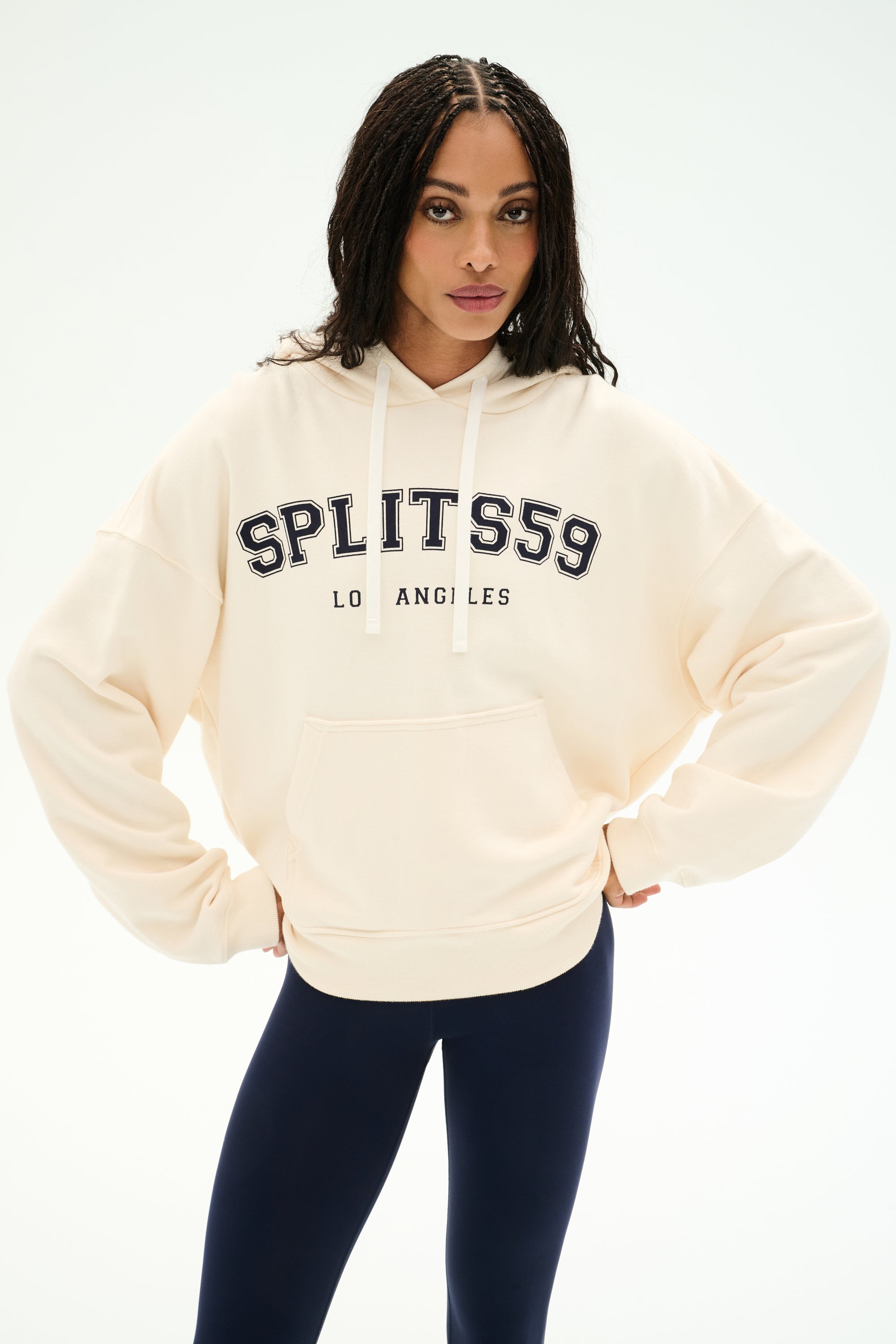 A person wearing the SPLITS59 Stella French Terry Oversized Hoodie in Creme/Splits59 Los Angeles, featuring soft cotton fabric, and navy leggings stands with hands on hips against a plain white background.