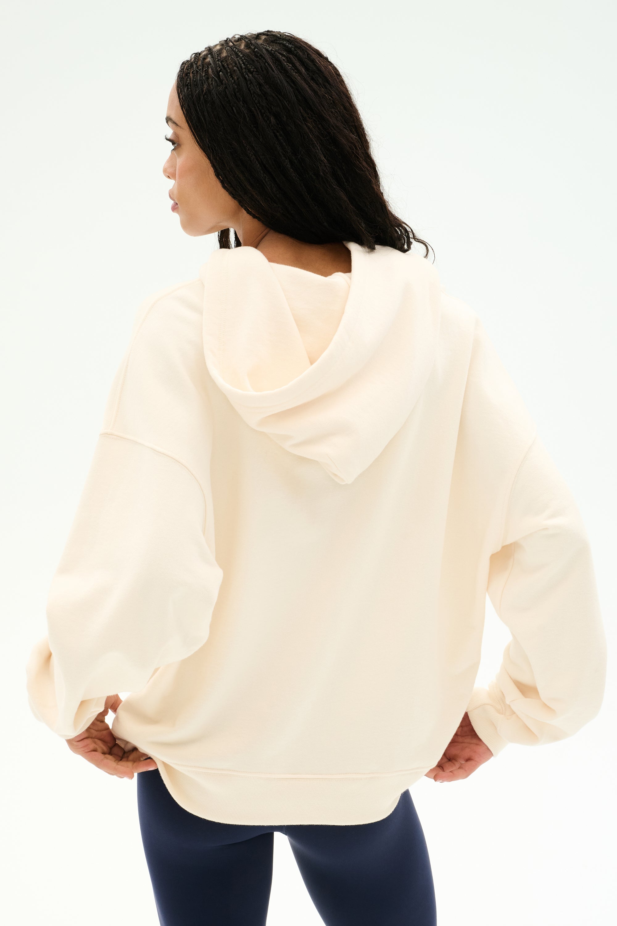 A woman stands with her back to the camera, hands on her hips, wearing the SPLITS59 Stella French Terry Oversized Hoodie in Creme, paired with navy leggings, set against a plain white background.