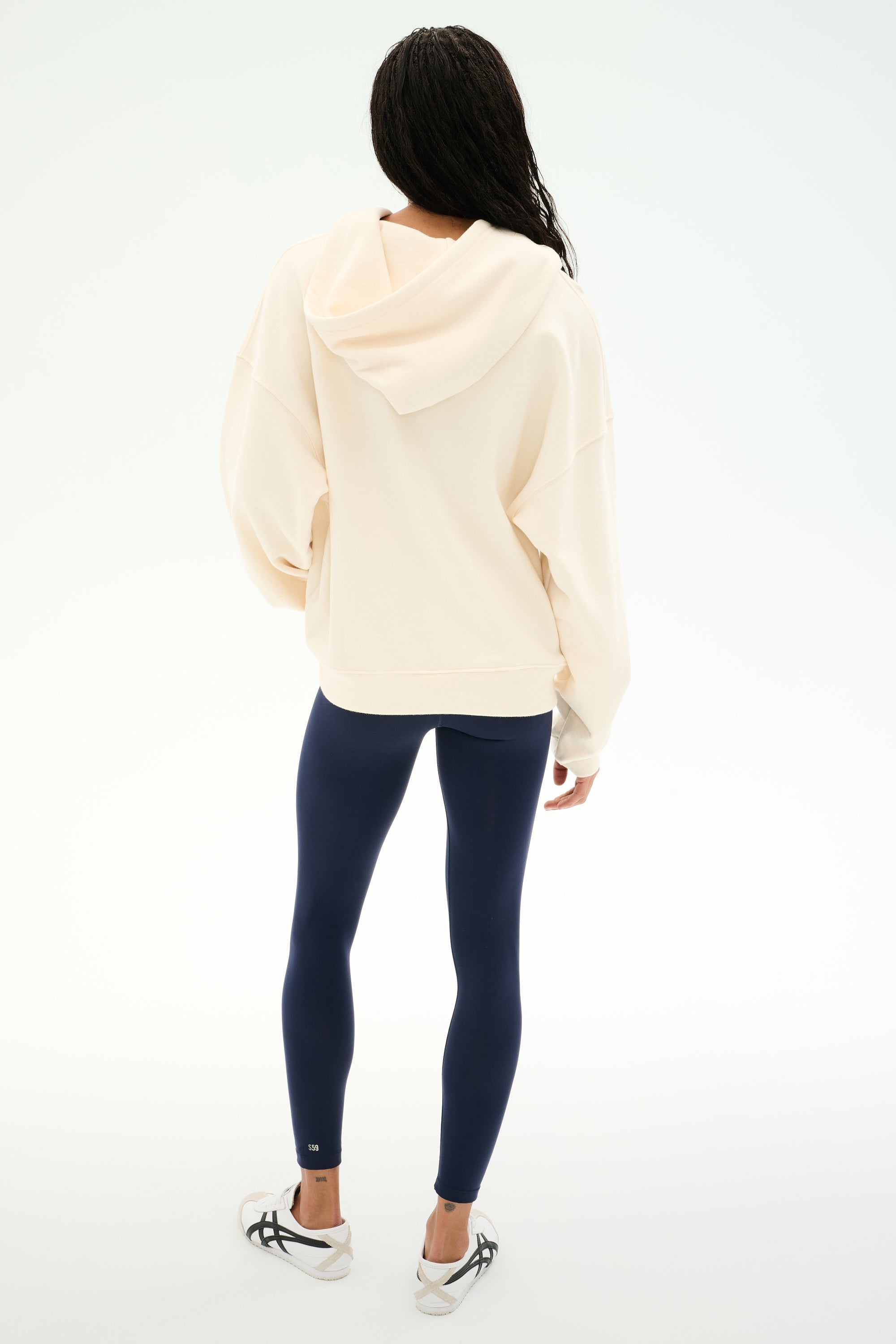 A person stands facing away against a plain white background, wearing the SPLITS59 Stella French Terry Oversized Hoodie in Creme, made from 100% cotton, paired with navy leggings and white athletic shoes.