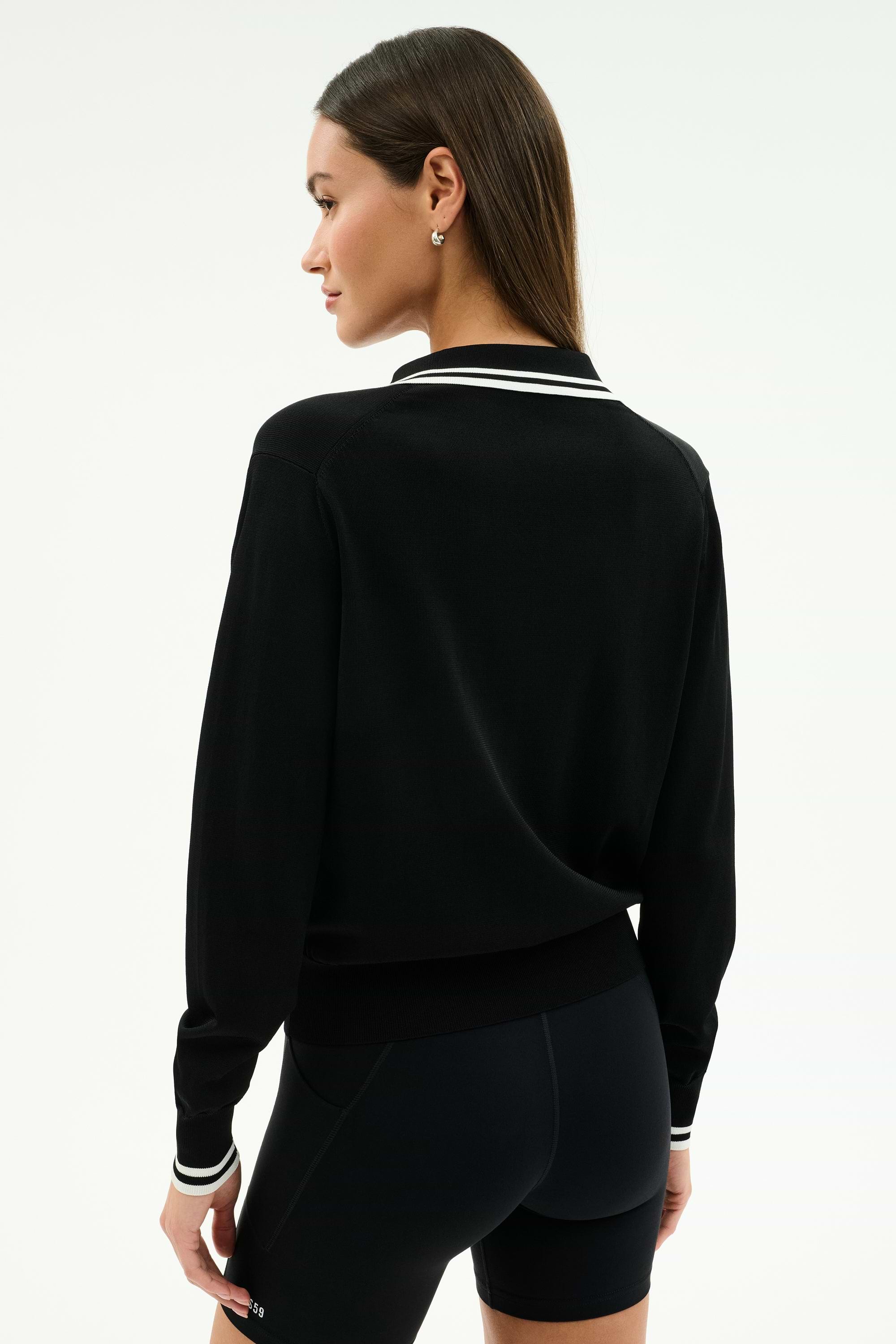 A woman stands with her back to the camera in the SPLITS59 Taylor Knit Polo in Black/White, a soft rayon-nylon blend long-sleeve top featuring white trim, styled with black fitted shorts against a plain light background.