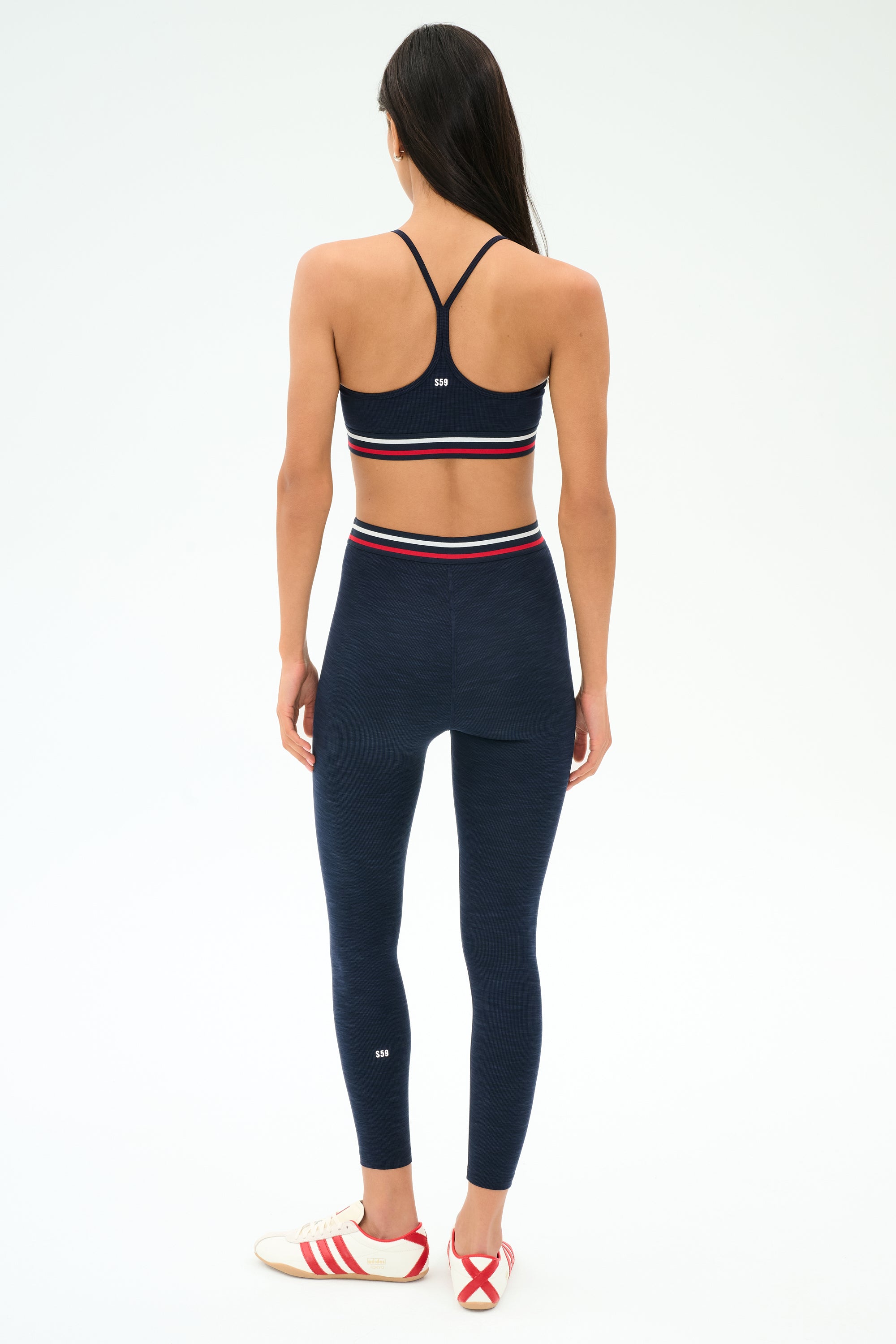 A woman faces away, wearing the SPLITS59 Cali Active Rib Bra in Heather Indigo with matching high-waisted leggings featuring red, white, and blue bands, paired with white sneakers with red stripes.