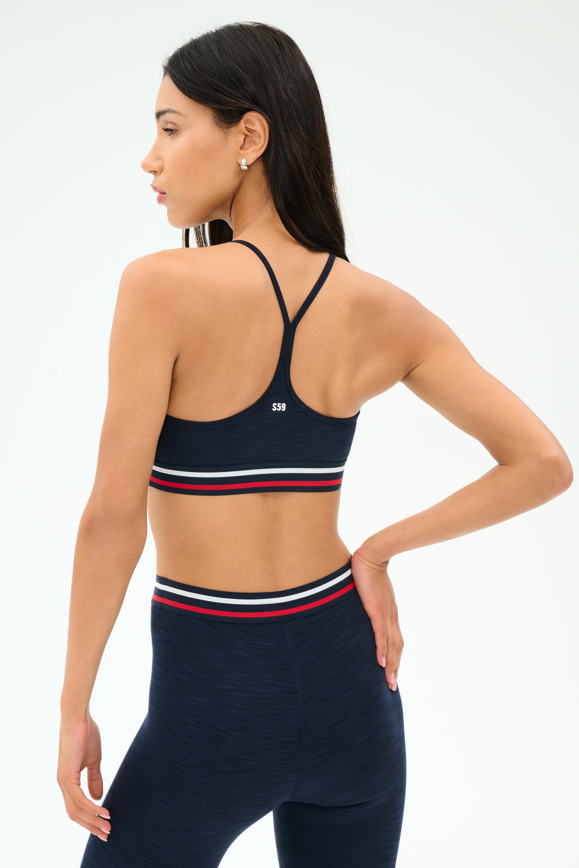 A woman poses from the back against a plain background, wearing the SPLITS59 Cali Active Rib Bra in Heather Indigo and matching high-waisted leggings with red, white, and navy striped bands.