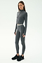 A woman wears the SPLITS59 Bailey Active Rib High Waist 7/8 leggings in Heather Grey with a fitted, long-sleeve top and black sneakers, standing against a plain white background.