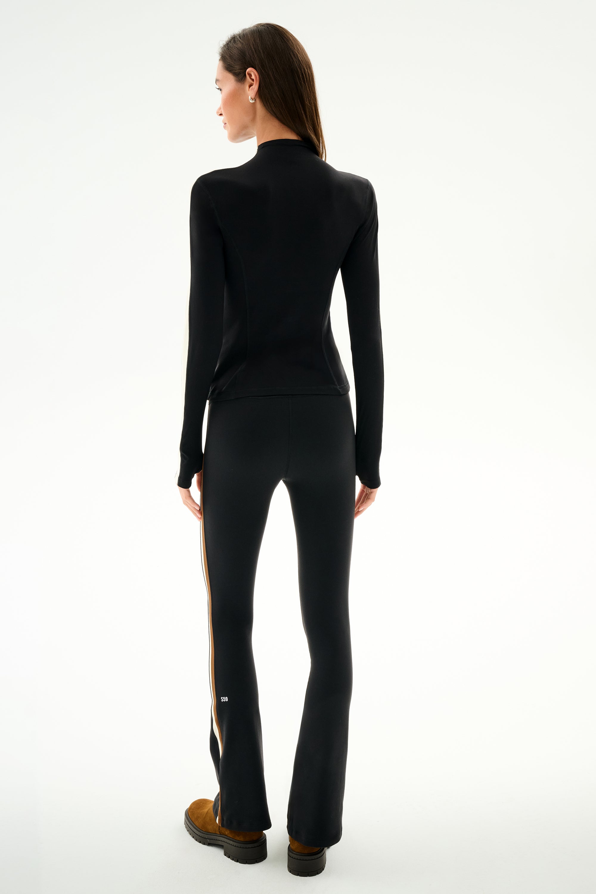 A person faces away wearing the SPLITS59 Isabelle Airweight Lite Mock Neck - Black/Creme, black side-striped pants, and brown shoes against a plain white background.