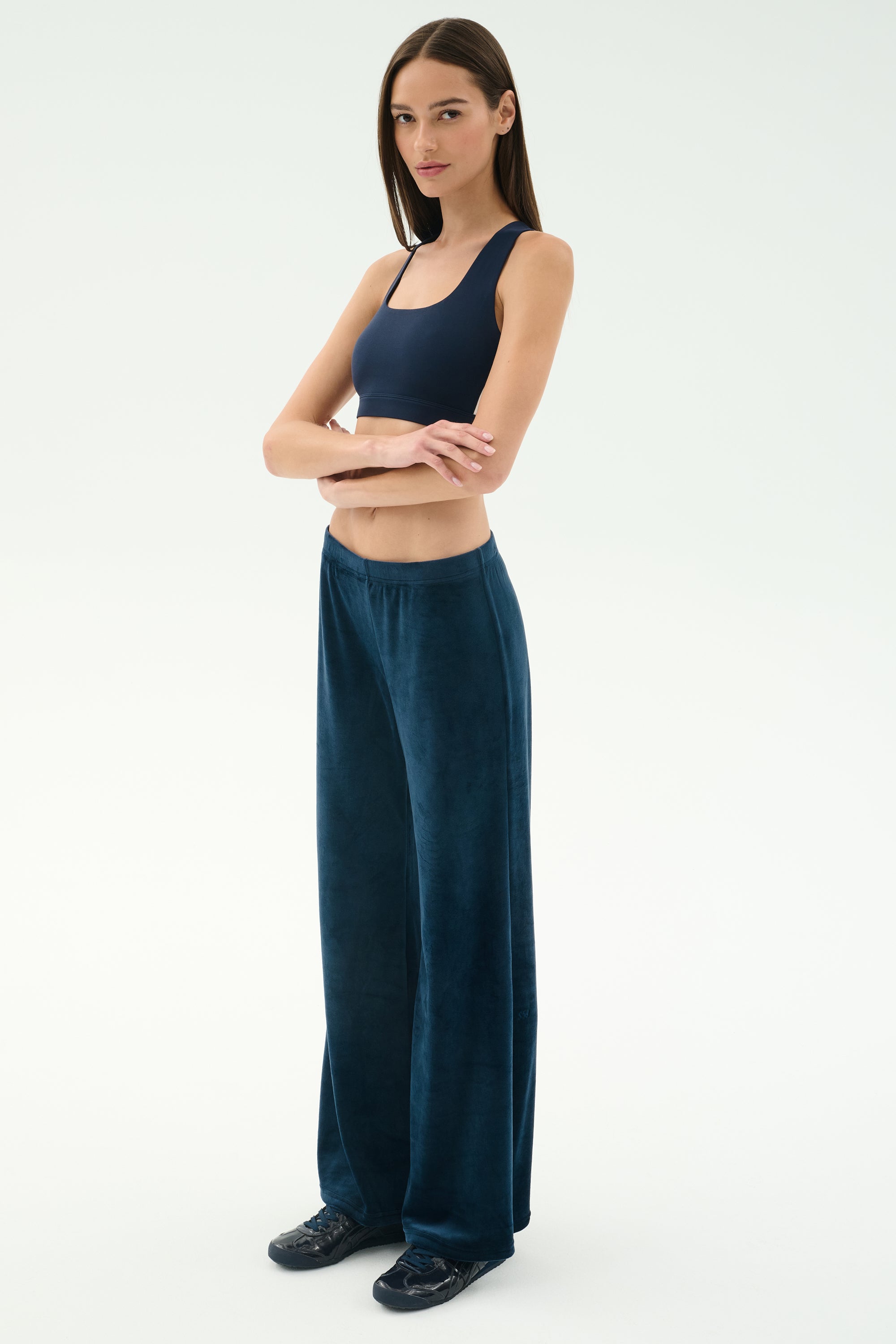 A woman stands with arms crossed, wearing the SPLITS59 Joy Velour Pant in Indigo and a navy crop top, paired with black shoes against a plain light background.