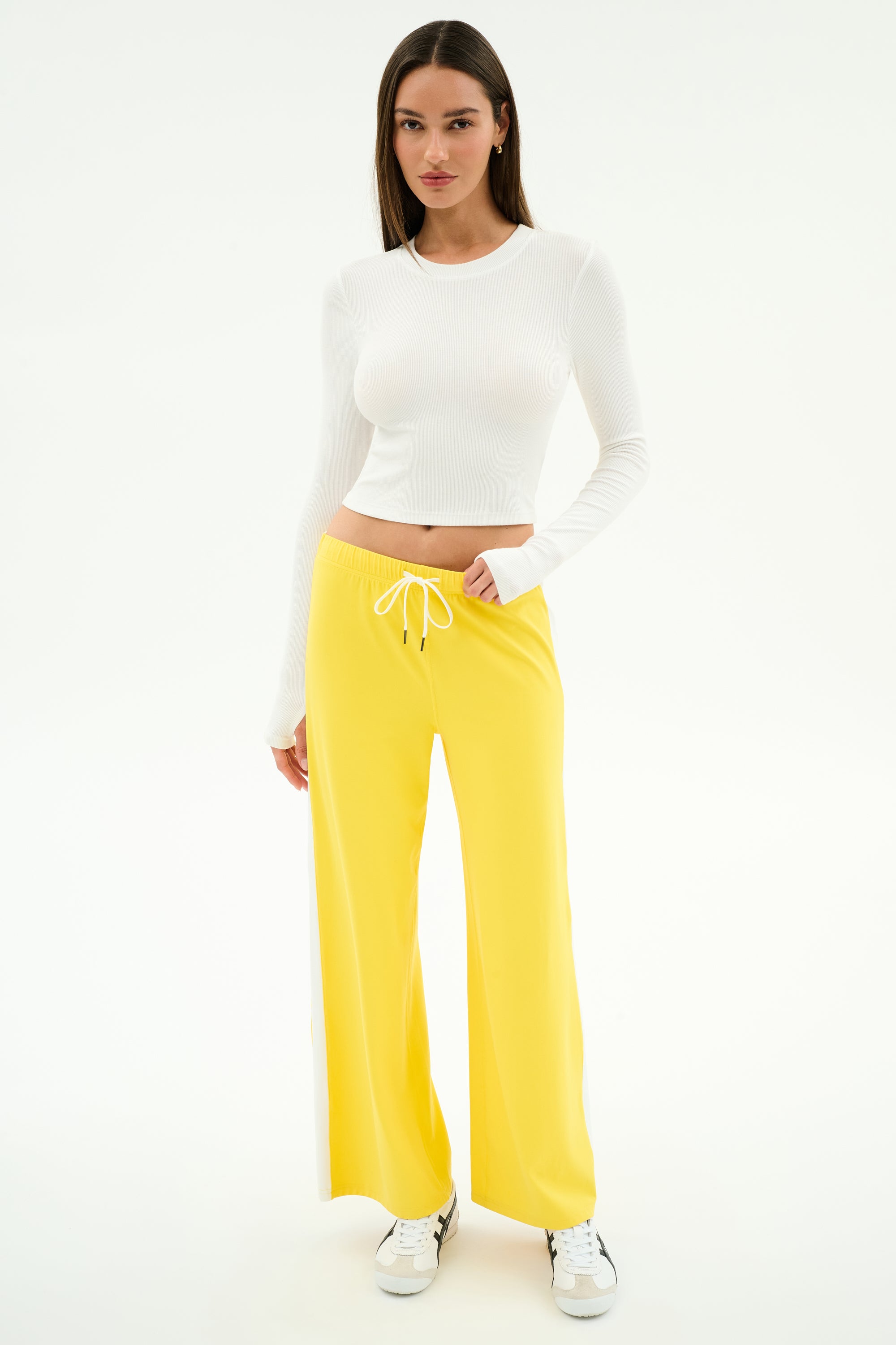 A woman stands in a studio wearing the SPLITS59 Louise Rib Long Sleeve Crop in white, paired with yellow wide-leg drawstring pants and white sneakers—an ideal gym-ready look—against a plain white background.