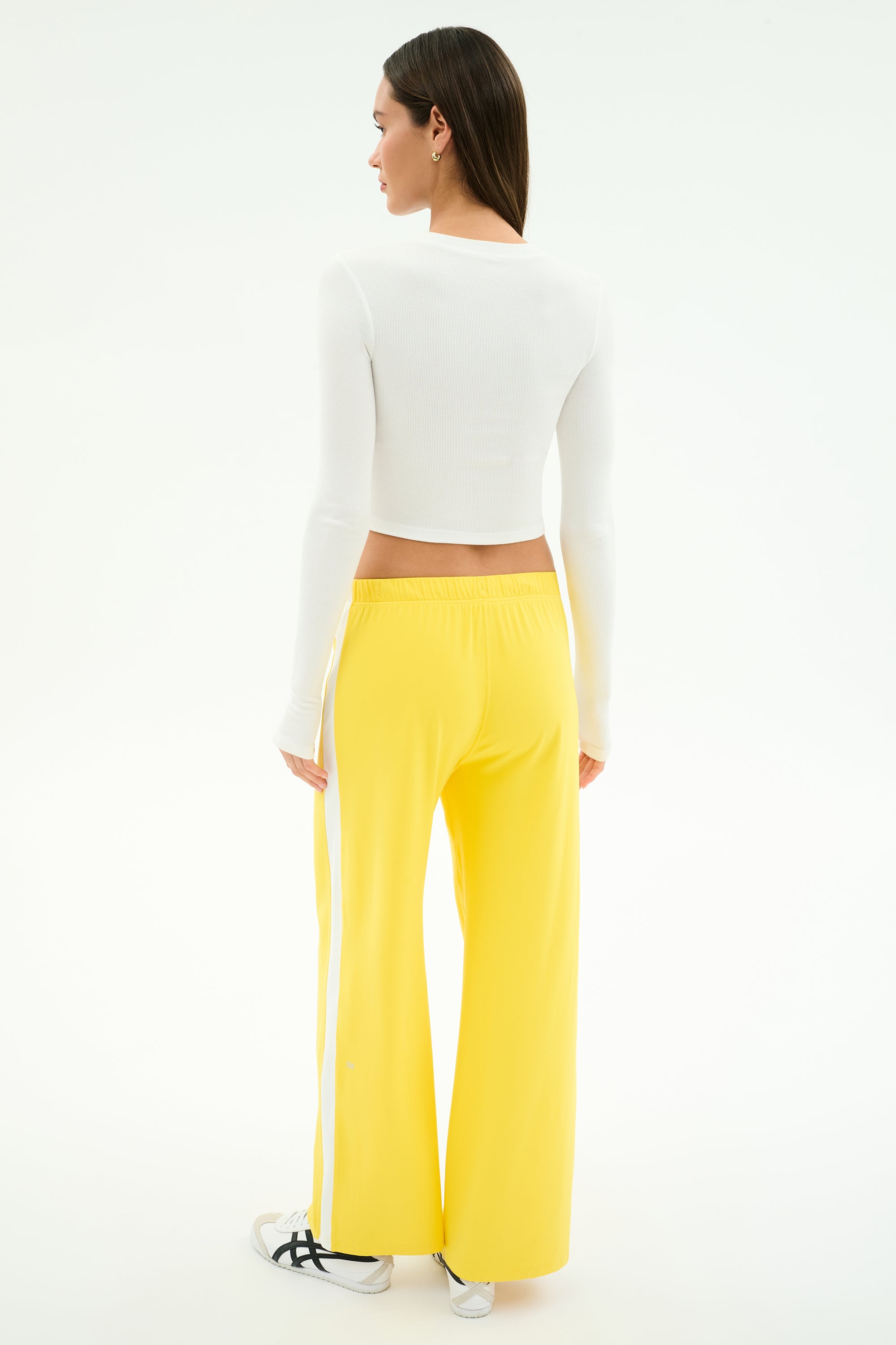 A person stands facing away, wearing the SPLITS59 Louise Rib Long Sleeve Crop in white with bright yellow wide-leg pants and white sneakers featuring black accents, posed against a plain light background. Perfect style for post-gym workouts.