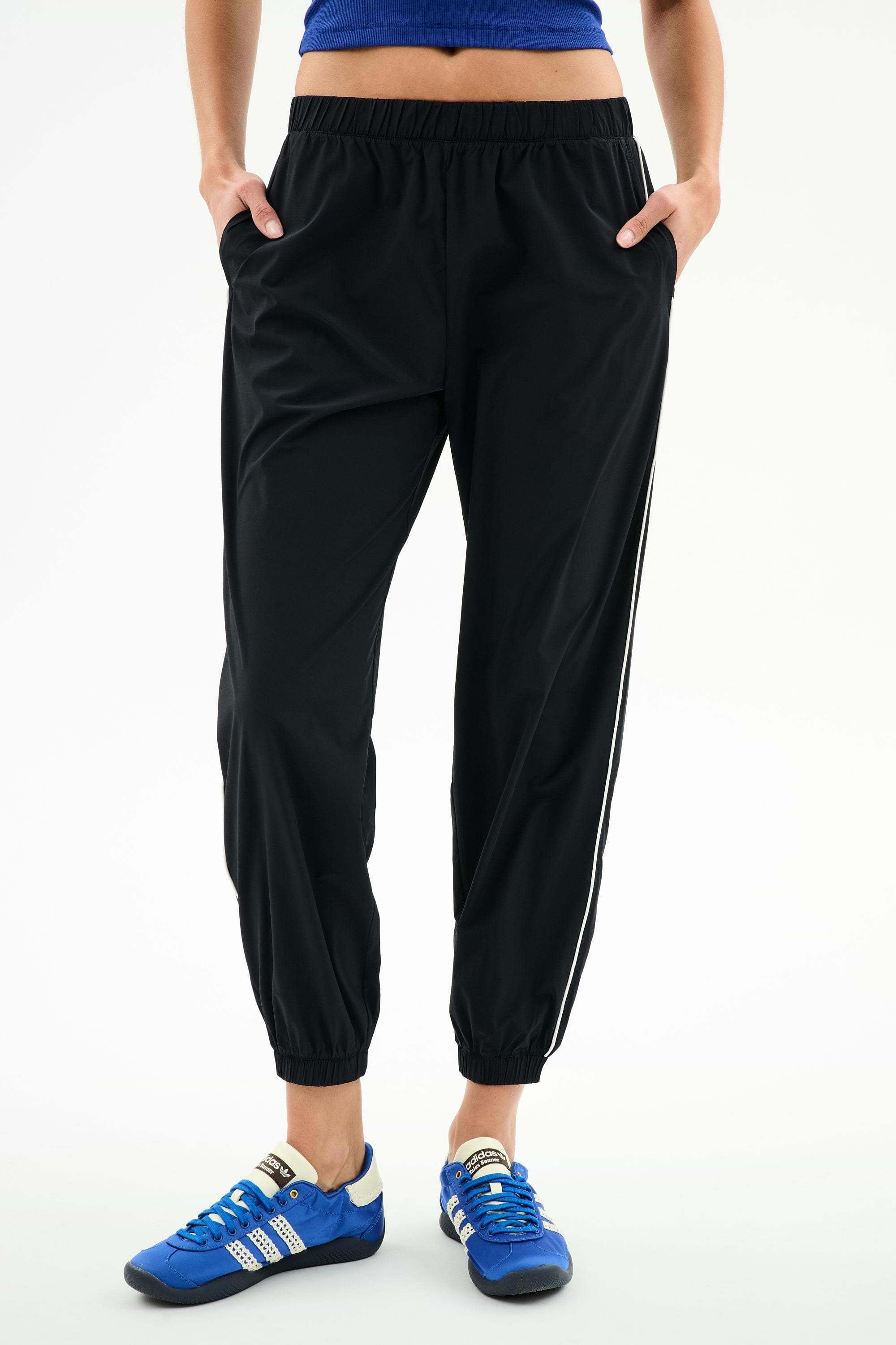 A person wears SPLITS59 Megan Woven Nylon Jogger w/ Piping - Black/Creme, a blue top, and blue athletic shoes, standing with hands in pockets against a plain white background.