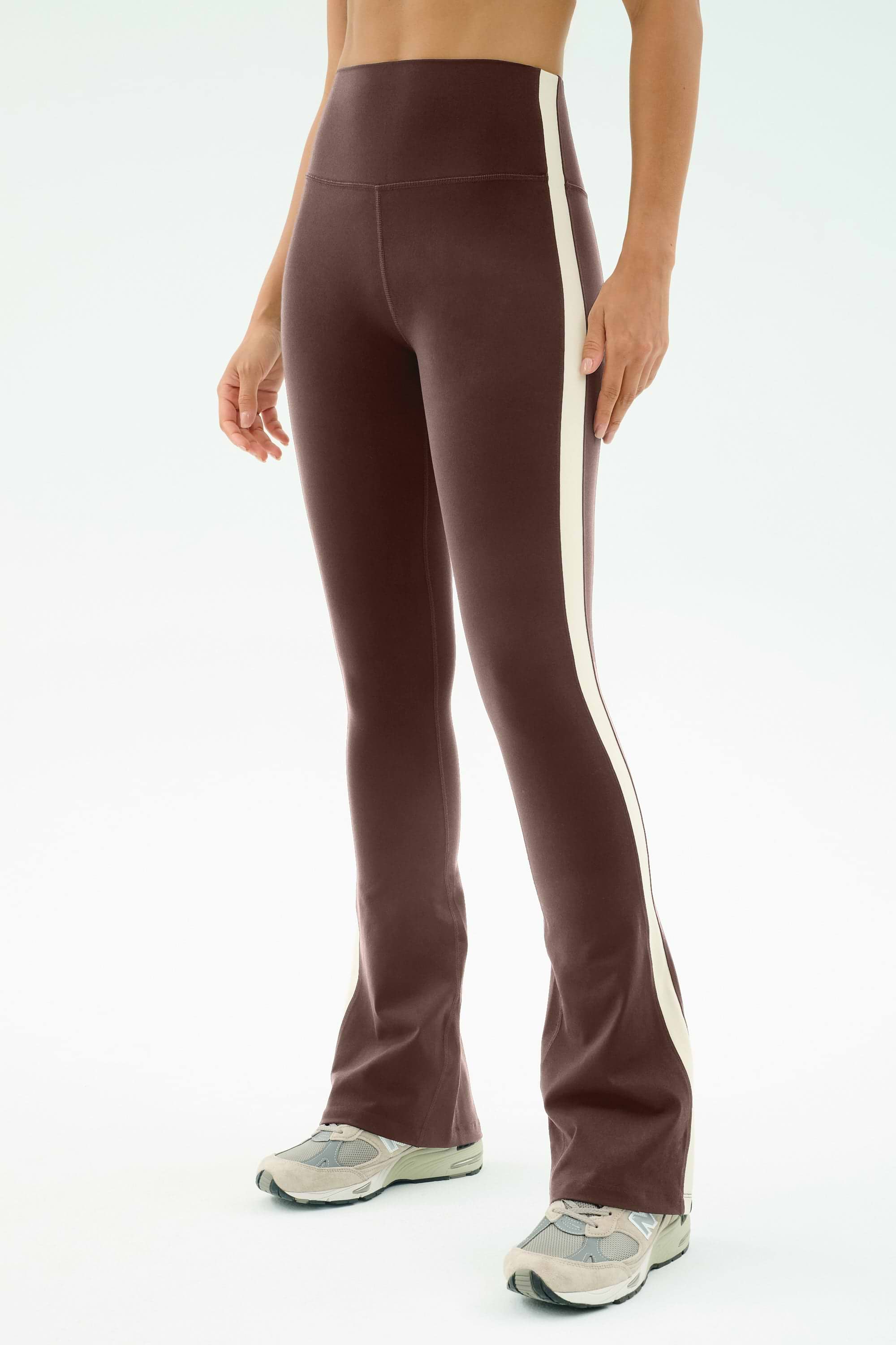 A person wears SPLITS59's Raquel High Waist Flared Legging in Dark Chocolate/Creme, featuring a comfy nylon-spandex blend and white side stripe, paired with gray sneakers against a light background.