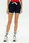 A person wears a red top, SPLITS59 Reena Sweatshort - Indigo, white striped socks, and white sneakers, standing against a plain background.