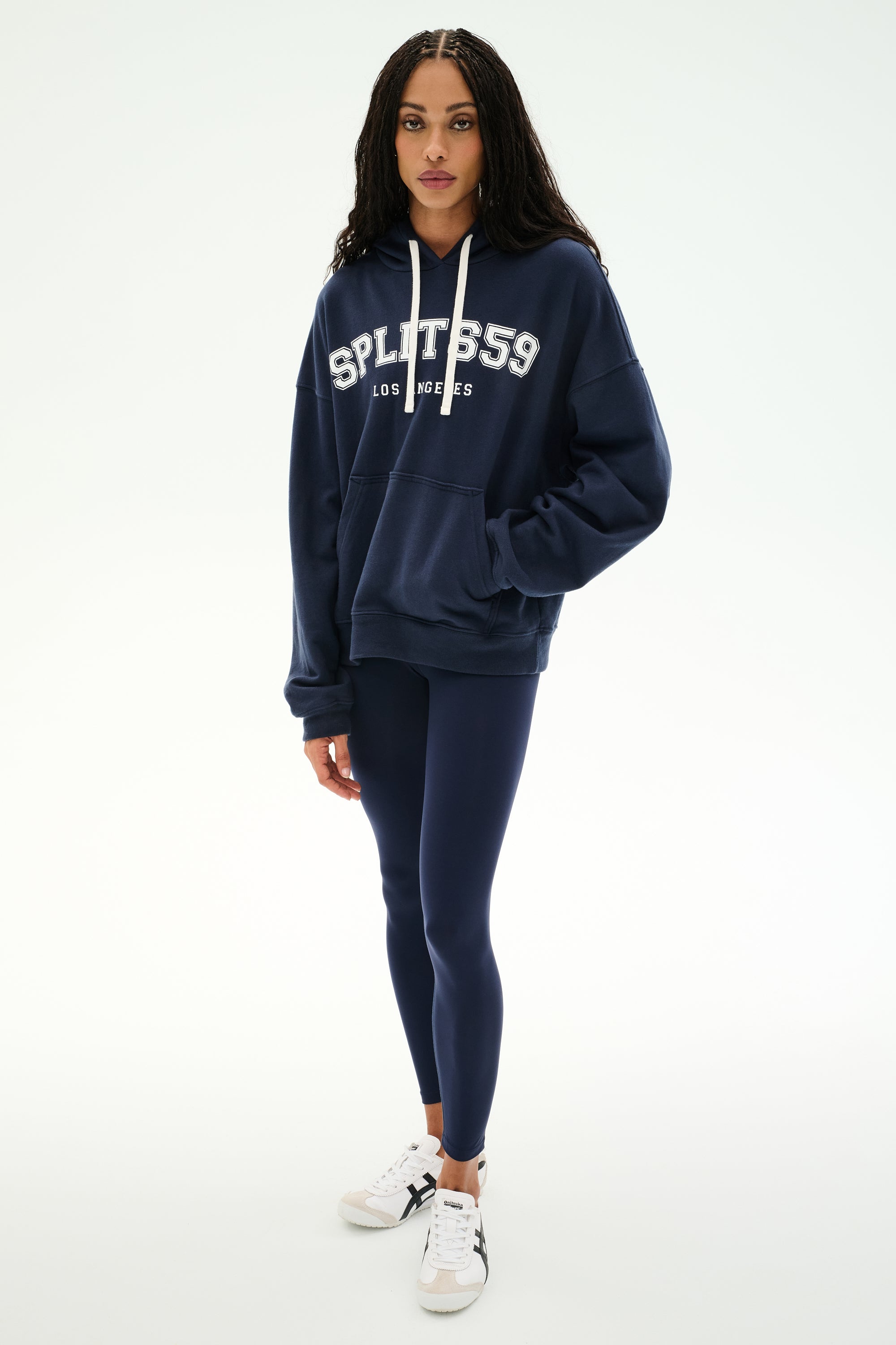 A woman stands facing forward, wearing the SPLITS59 Stella French Terry Oversized Hoodie in Indigo/Splits59 Los Angeles, crafted from soft cotton fabric. She pairs it with navy leggings and white sneakers against a plain white background.